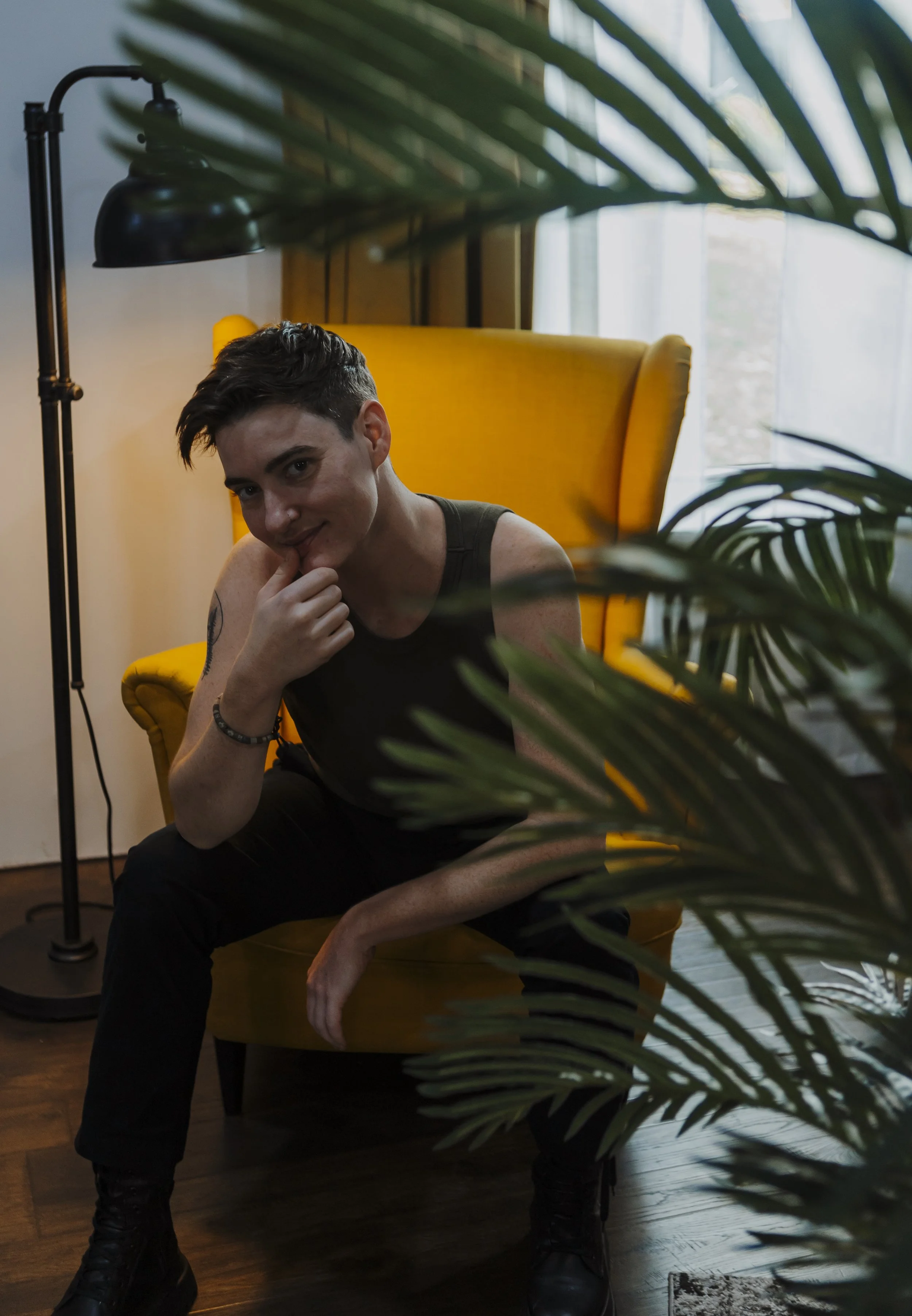 A young person with short dark hair sitting on a yellow chair in a room, smiling and touching their chin with one hand, partly obscured by large green plant leaves.