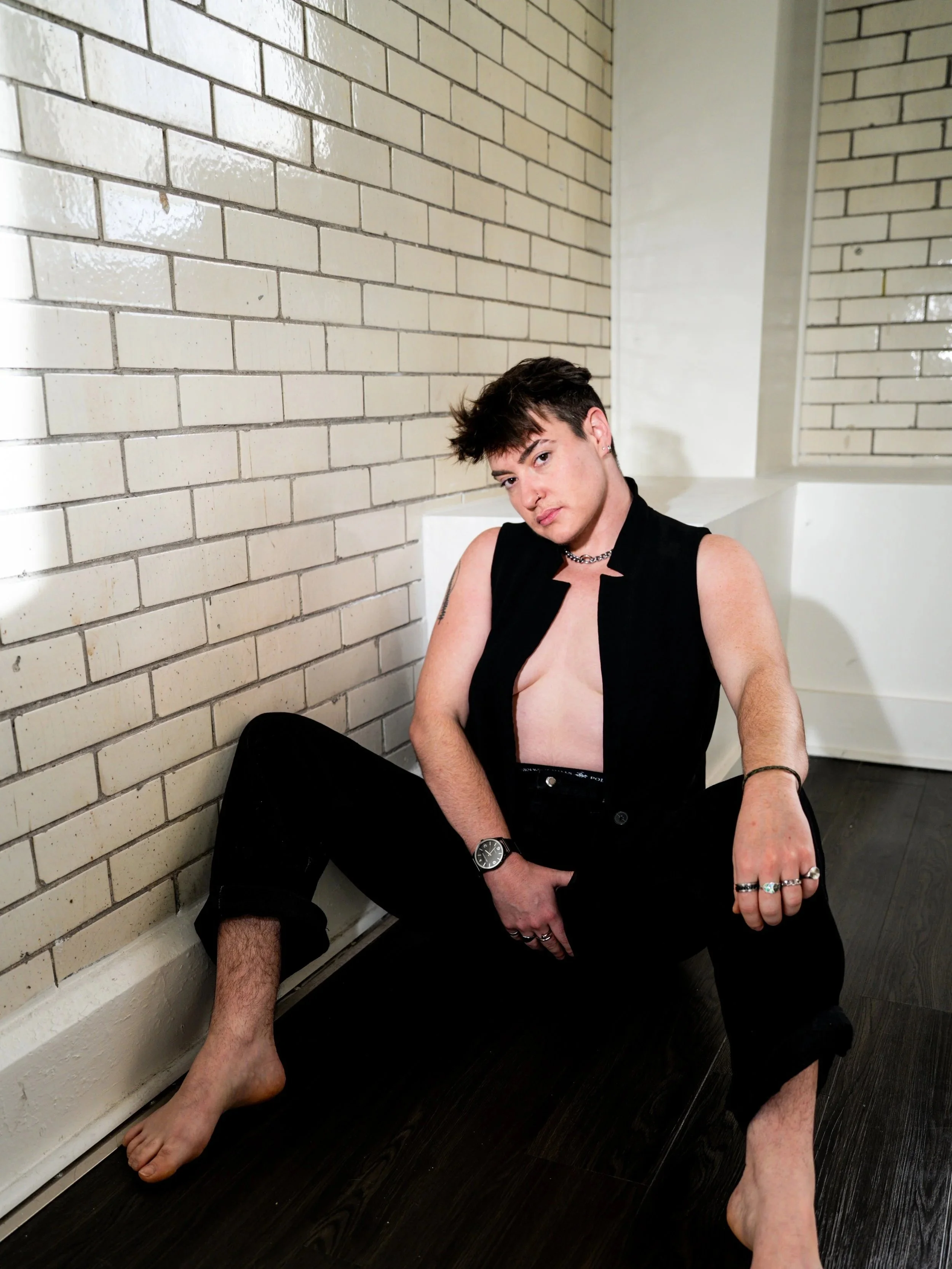A person with short dark hair sitting on the floor with legs apart, wearing black pants, a sleeveless black vest, multiple rings, a wristwatch, and a necklace, posing against a white brick wall in a room with dark hardwood flooring.
