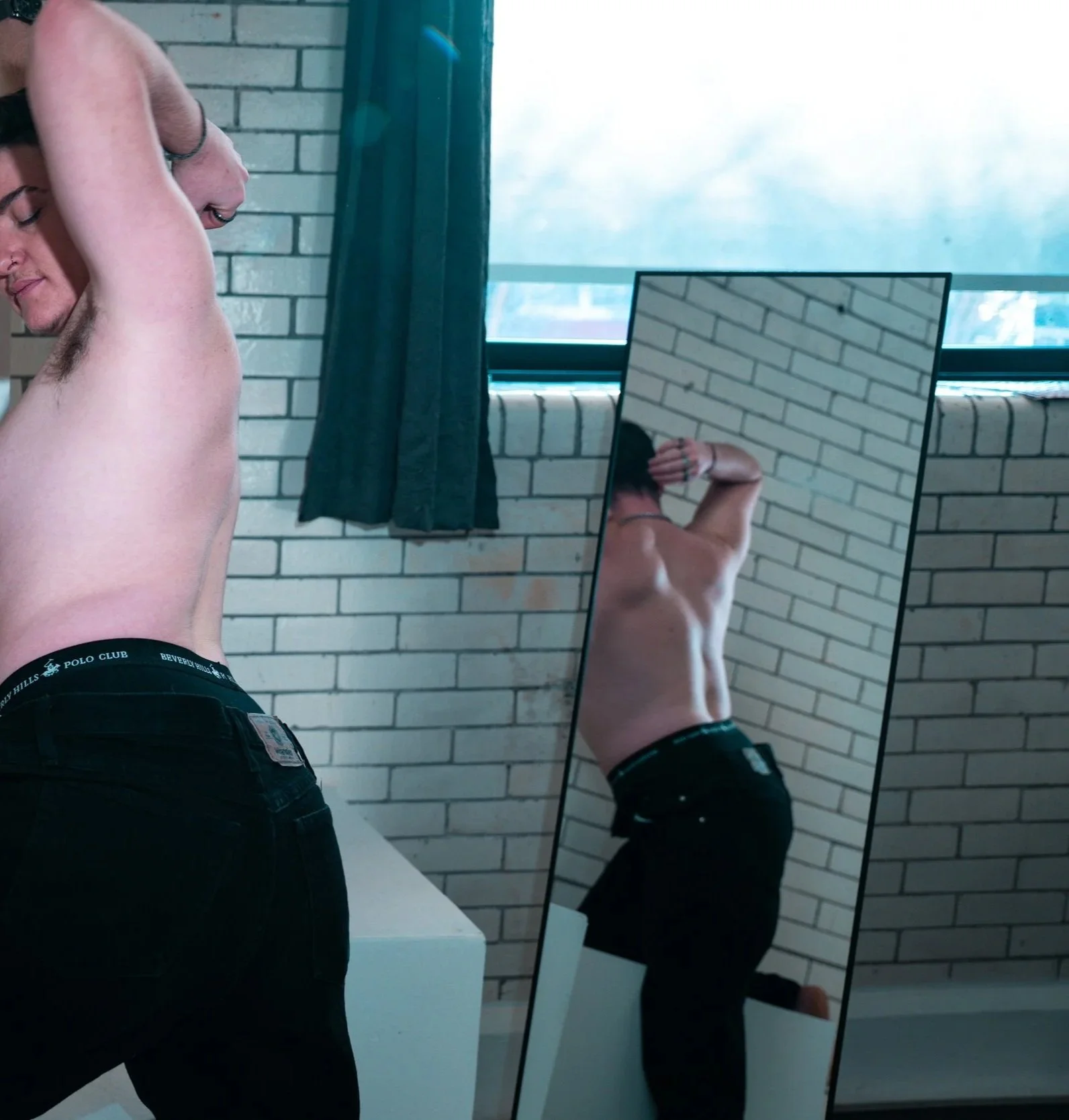A shirtless man with black jeans looking into a mirror, with his back turned, in an indoor setting with a brick wall and a window with dark curtains.