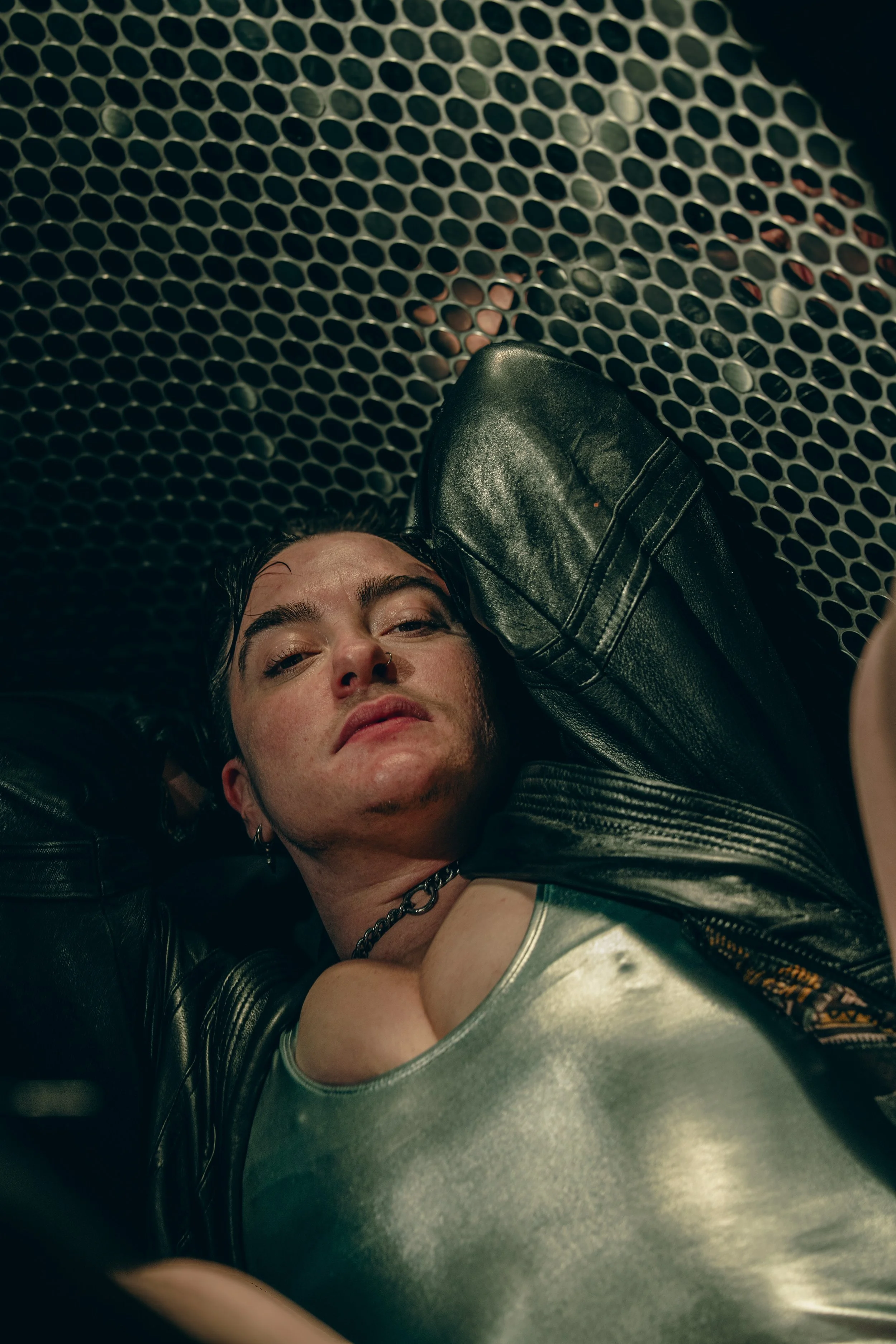 A person with short dark hair and light skin is lying down on a surface with a black leather jacket on, looking at the camera with a relaxed expression. The background features a perforated metallic surface with circular holes.