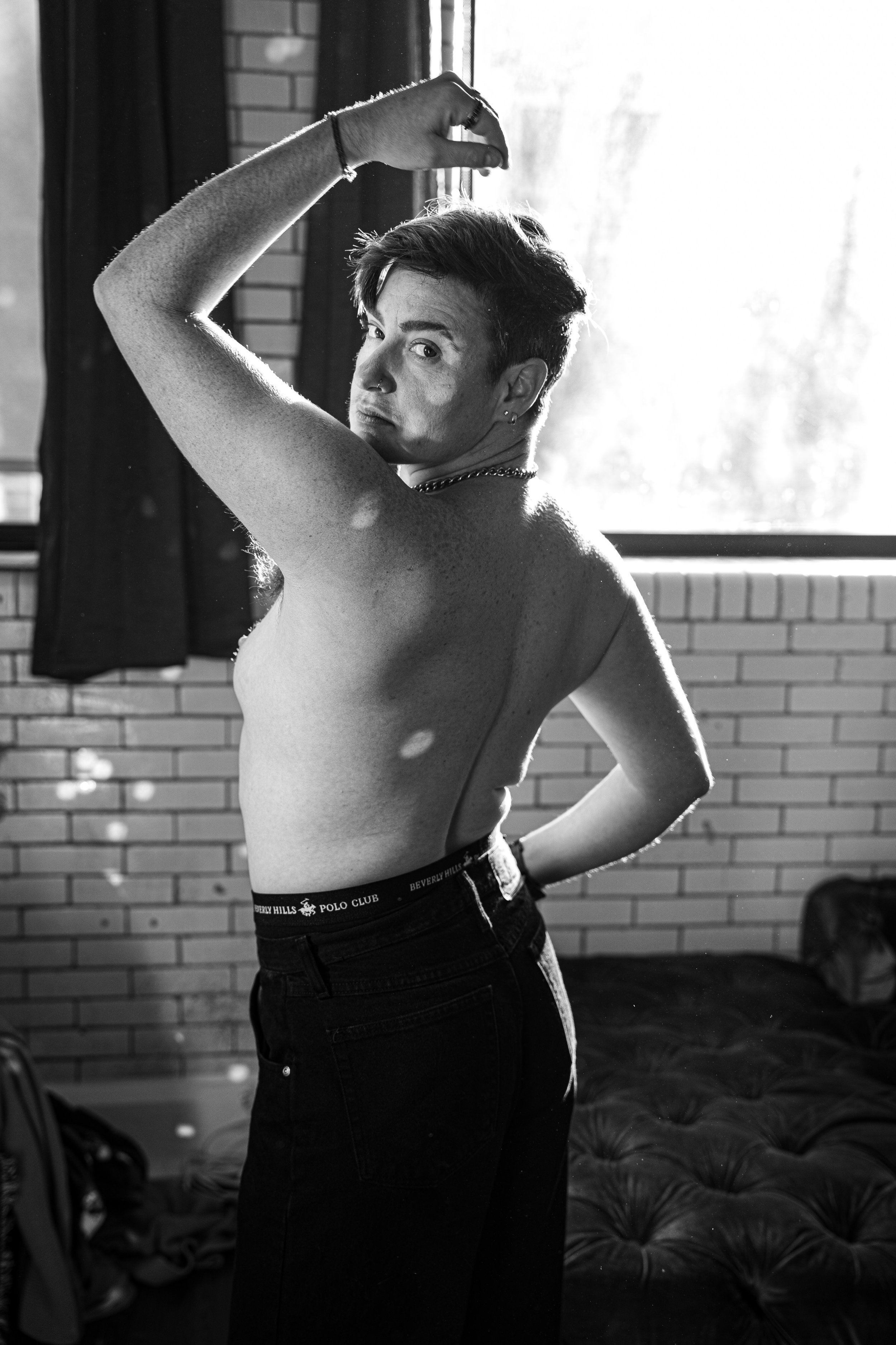 A person with short hair, tattoos, and jewelry, standing topless in a sunlit room with a brick wall and a bed in the background, looking over their shoulder at the camera.
