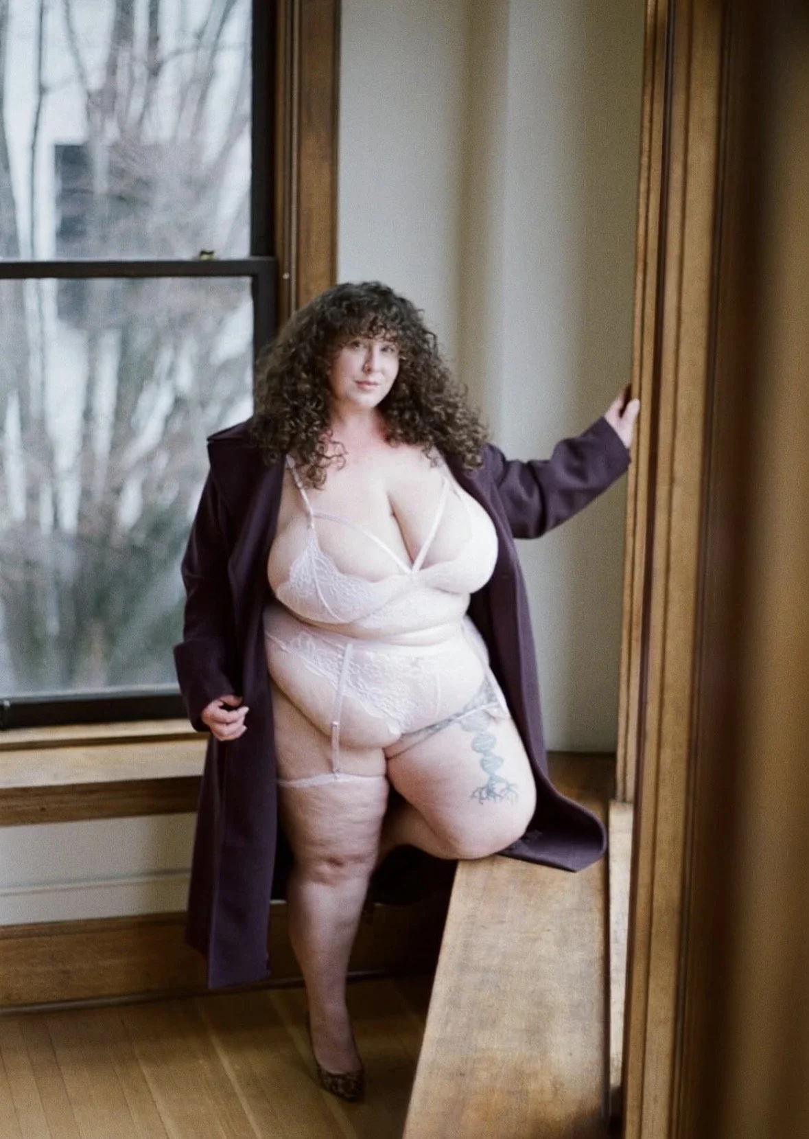 A woman with curly hair wearing lace lingerie and a dark coat, standing by a window in a room with wooden trim, during daylight.
