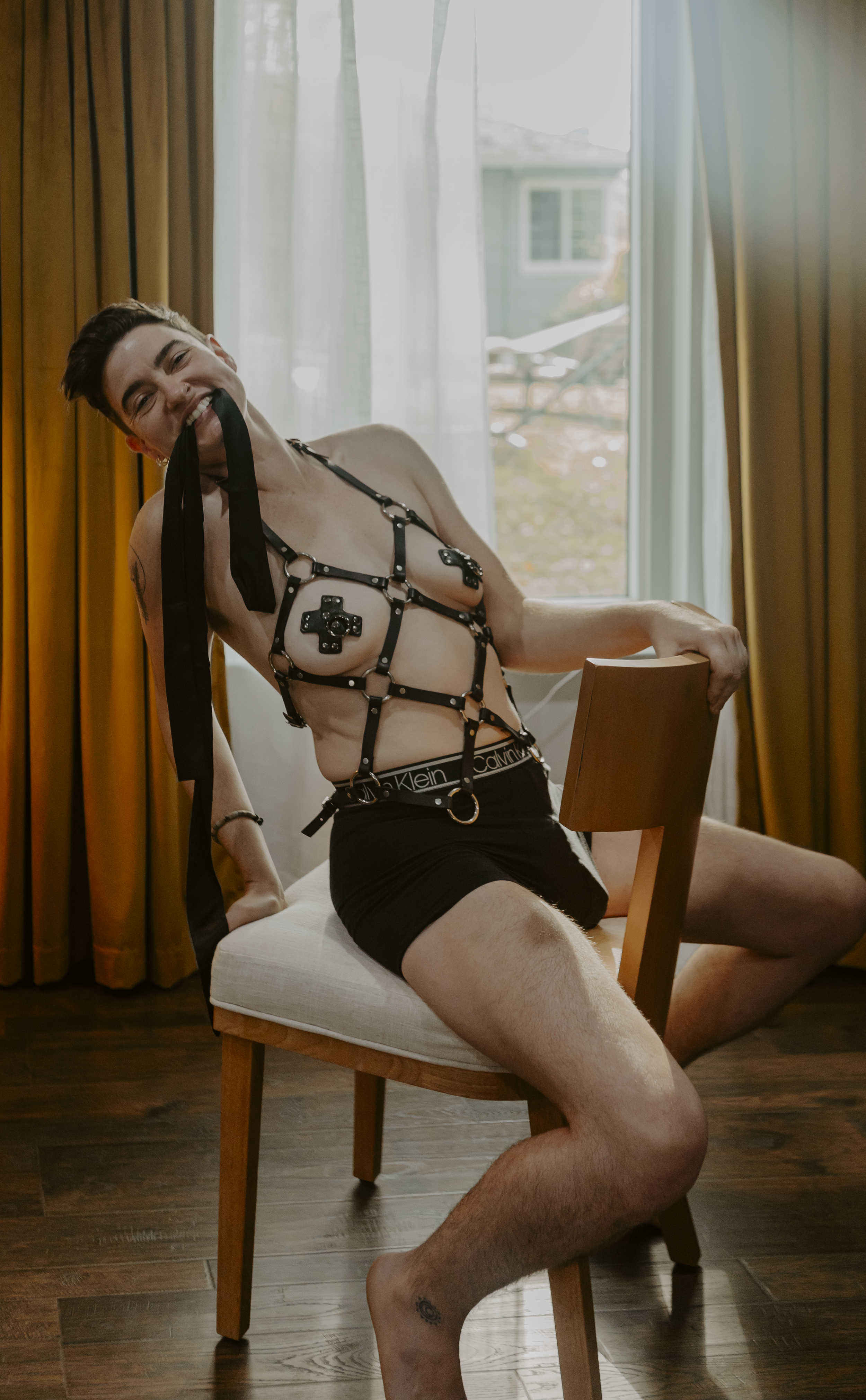 Person kneeling on the floor with their back to the camera, wearing black strappy underwear, with hands behind their head, in a room with dark walls, a mirror, and a red couch.