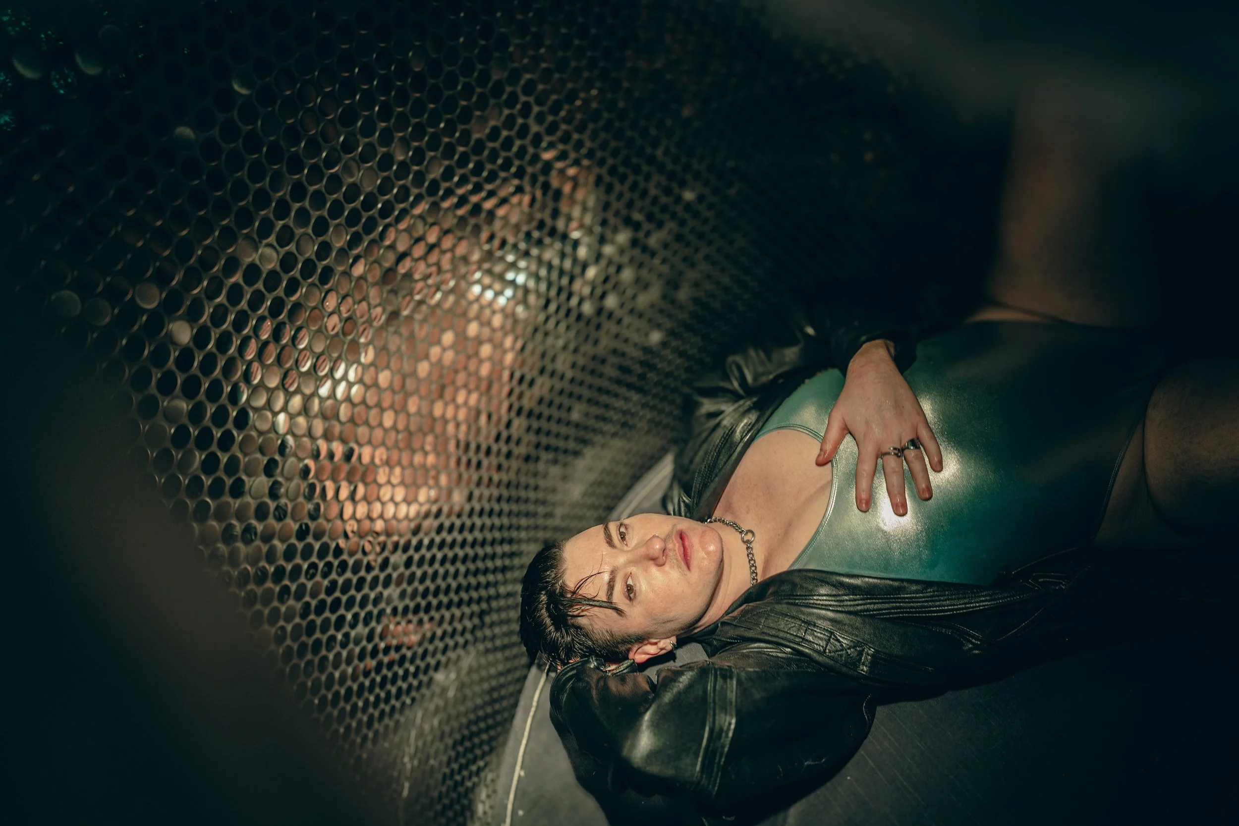 A woman with short dark hair and tattoos on her shoulder, lying on the ground inside a dark, industrial-style tunnel with a perforated metal wall, looking directly at the camera.