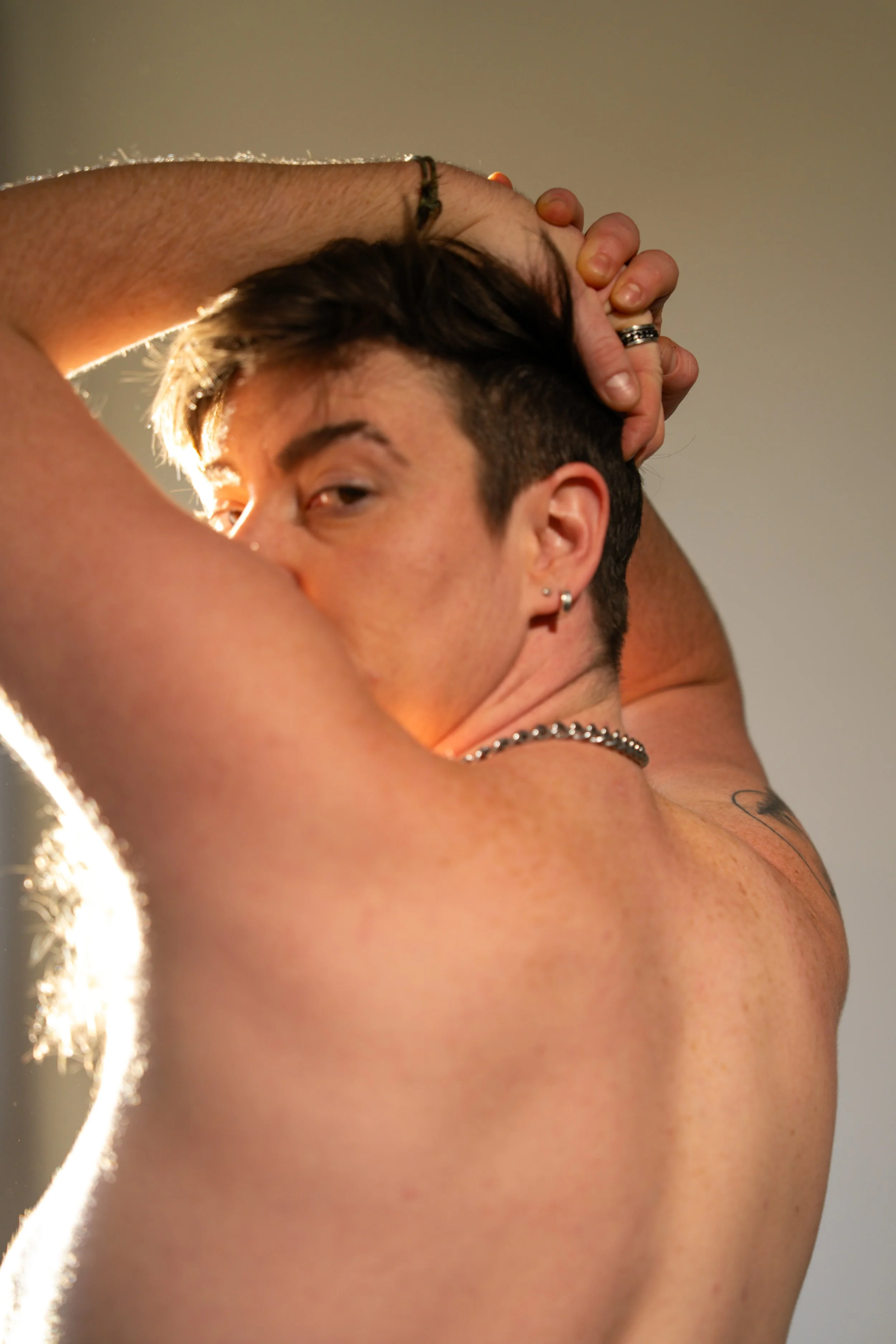 Close-up of a shirtless person with short dark hair, multiple earrings, wearing layered necklaces, holding their hair back with both hands, with a neutral background and warm lighting.