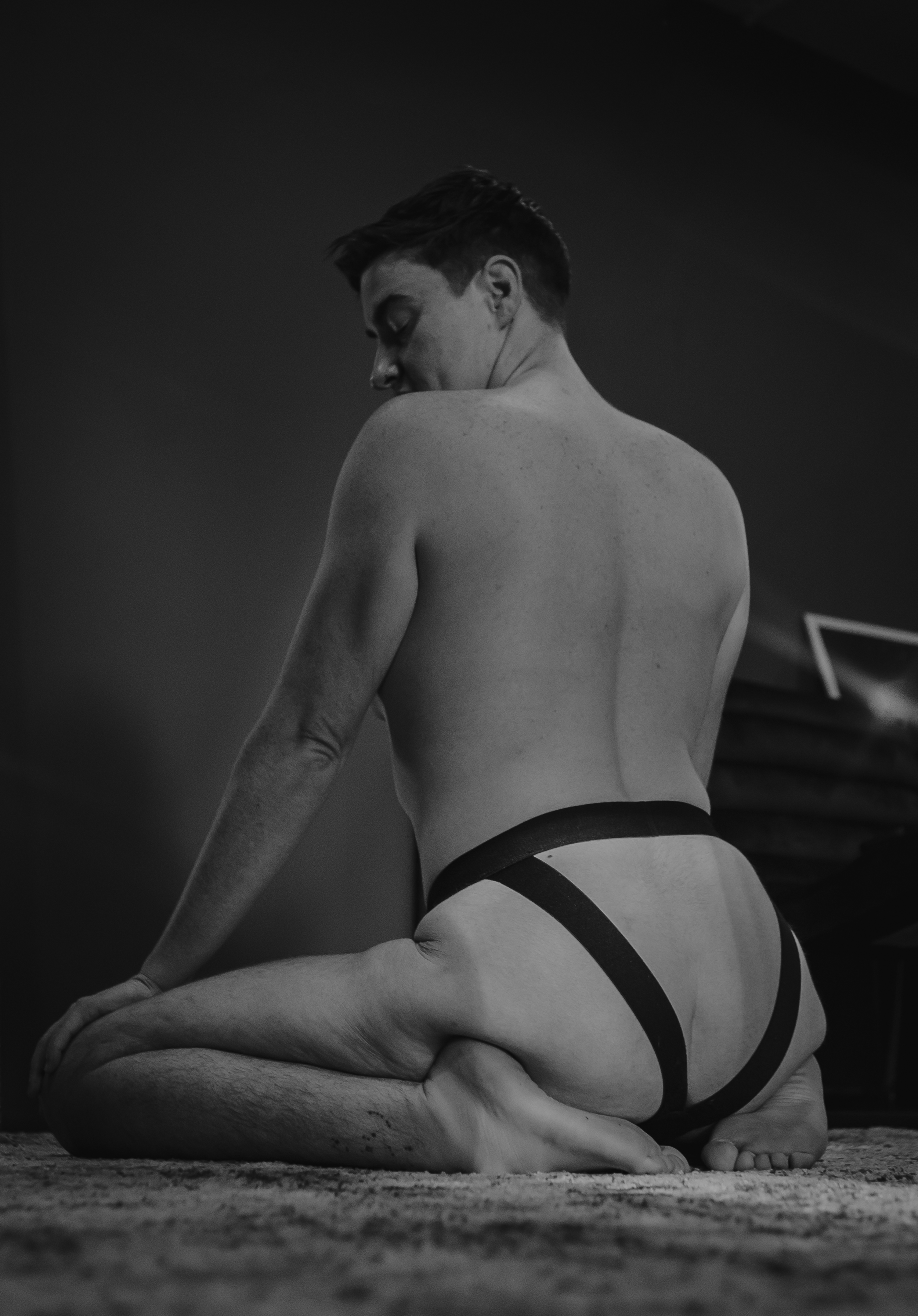 A black-and-white photo of a shirtless man kneeling on the floor with his head tilted down, wearing a harness-style strap around his waist, on a textured carpet in a dimly lit room.