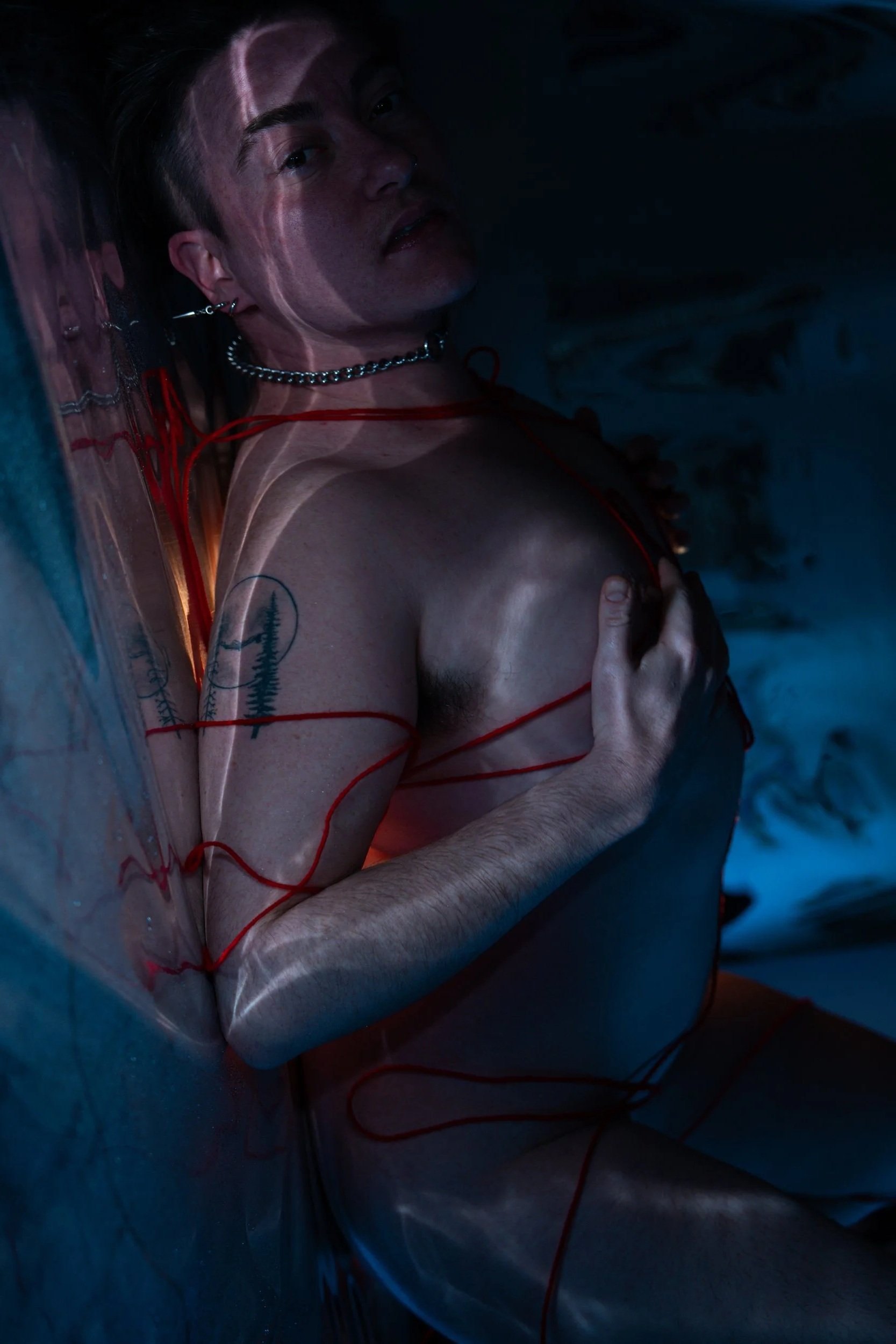 A young man lying on the ground with multiple red wires connected to his body, wearing earrings, a chain necklace, and a red strap, with tattoos on his arm, in a dark setting.