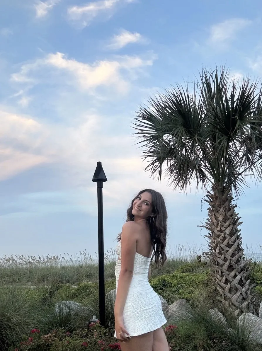A young woman in a white dress smiling and posing outdoors near a palm tree with a blue sky and clouds in the background.