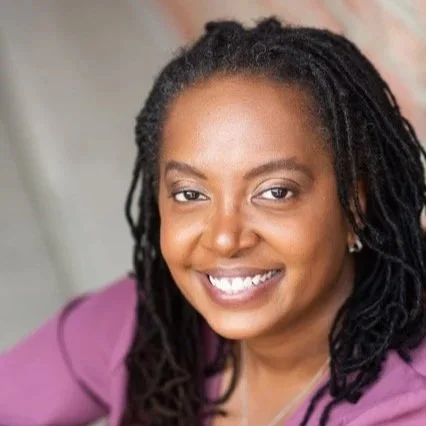Close-up portrait of an African American woman with long braided hair, smiling and wearing a purple top.