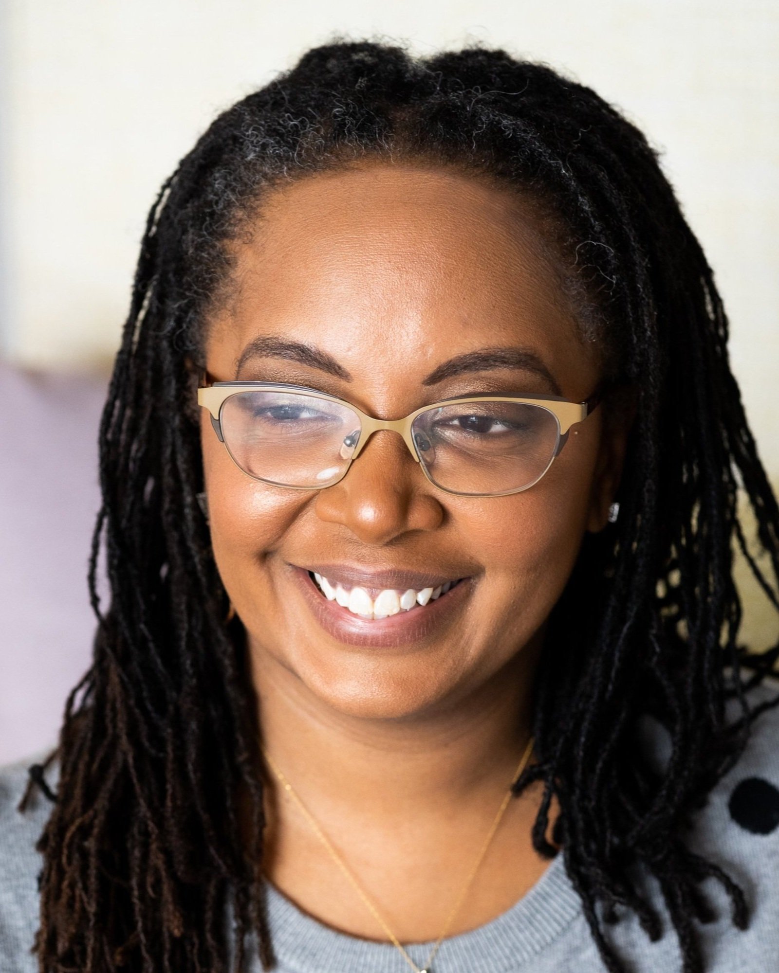 A smiling woman wearing glasses and a gray sweater with a polka dot pattern. She has long, black locs and is looking off to the side.