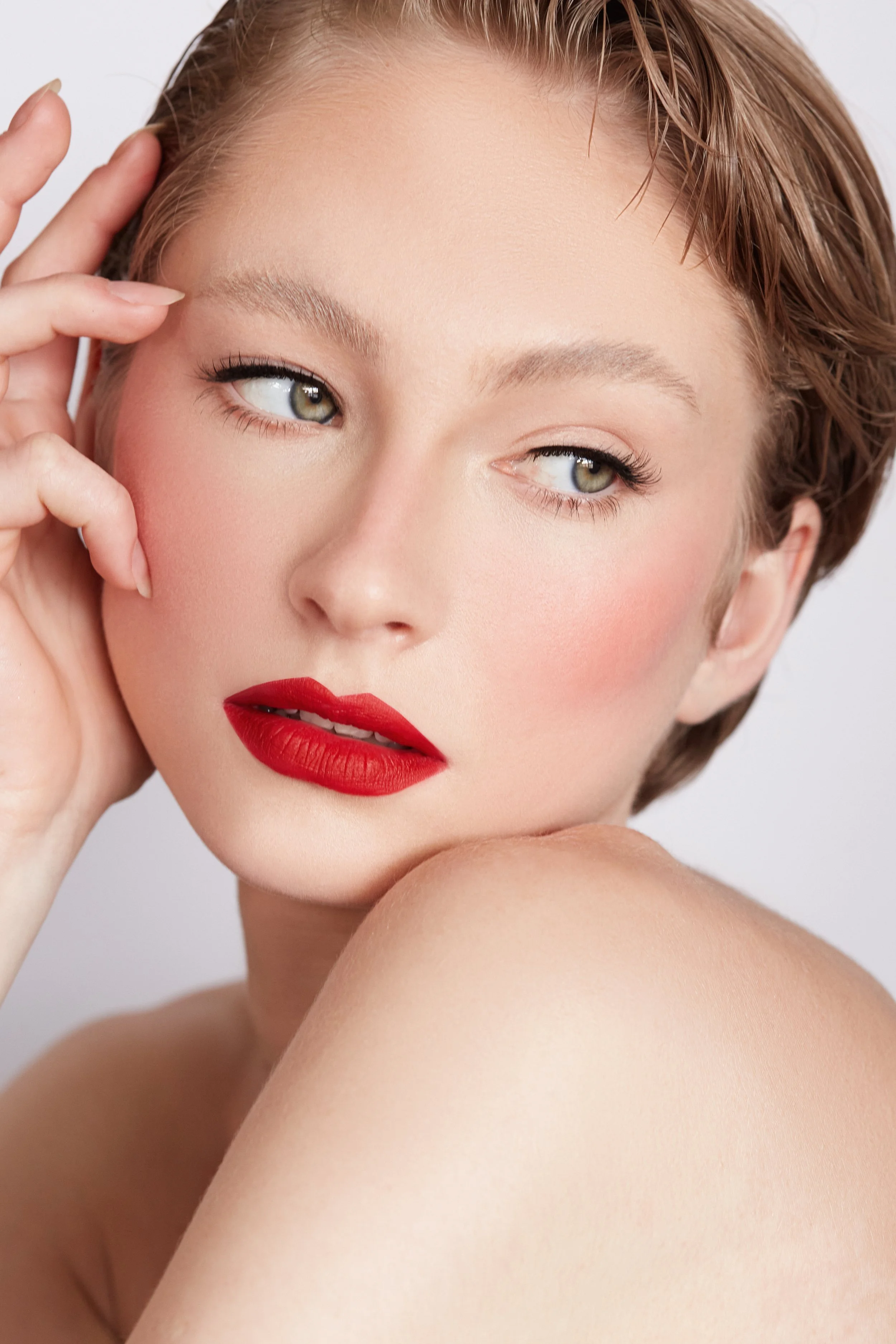 Close-up of a woman with fair skin, short brown hair, red lipstick, and natural makeup, posing with her hand near her face.