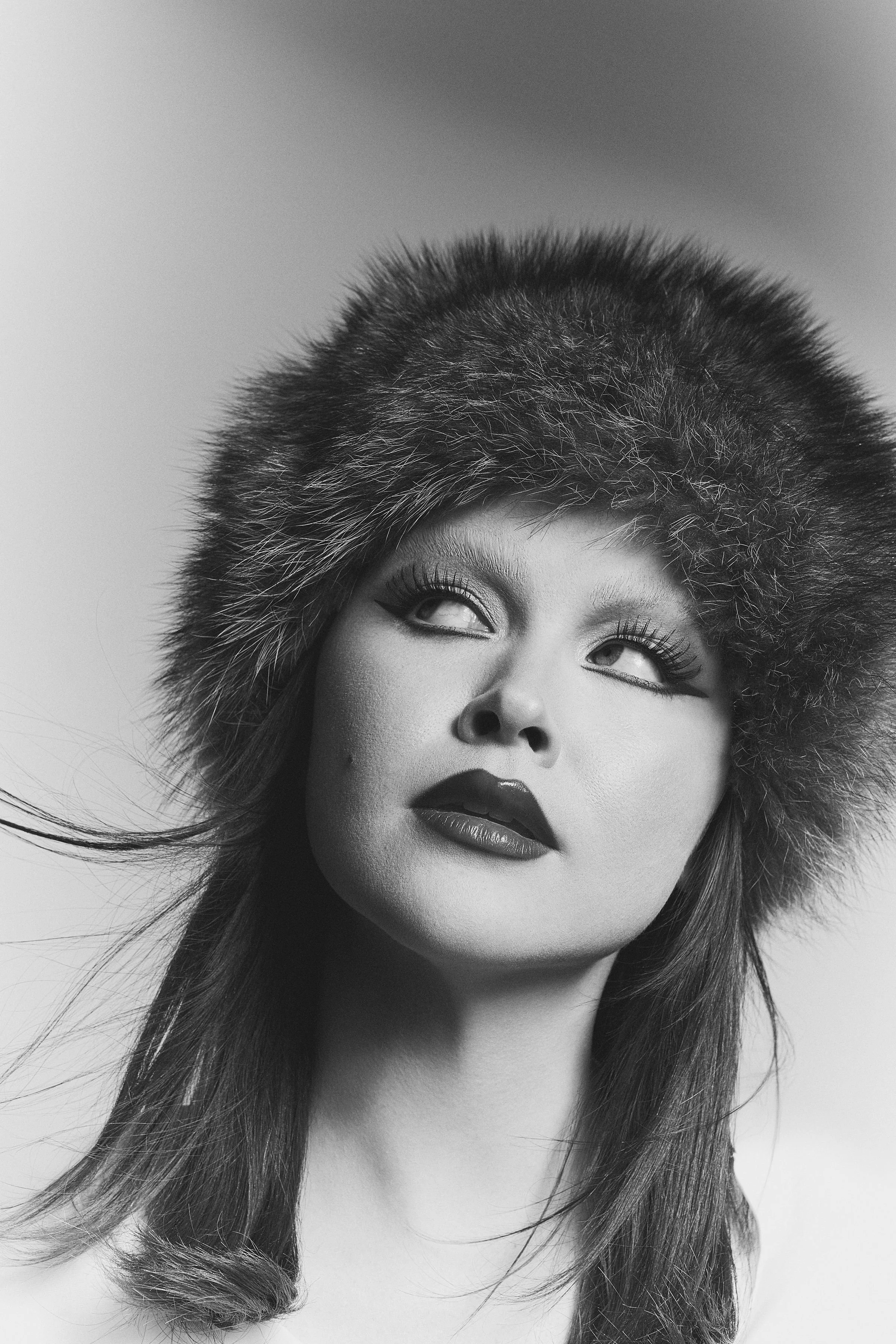 Black and white portrait of a woman wearing a large furry hat, with makeup emphasizing her eyes and lips, looking upward to the side.
