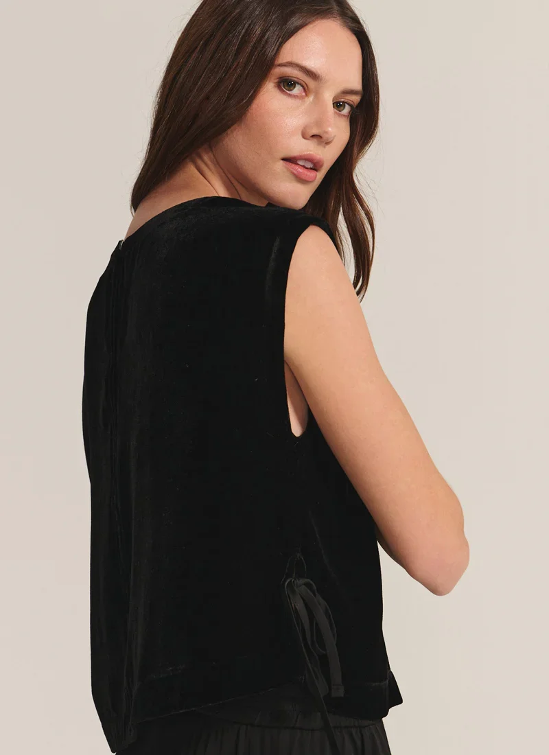 A woman with long brown hair looking over her shoulder at the camera, wearing a sleeveless black top, against a plain light background.