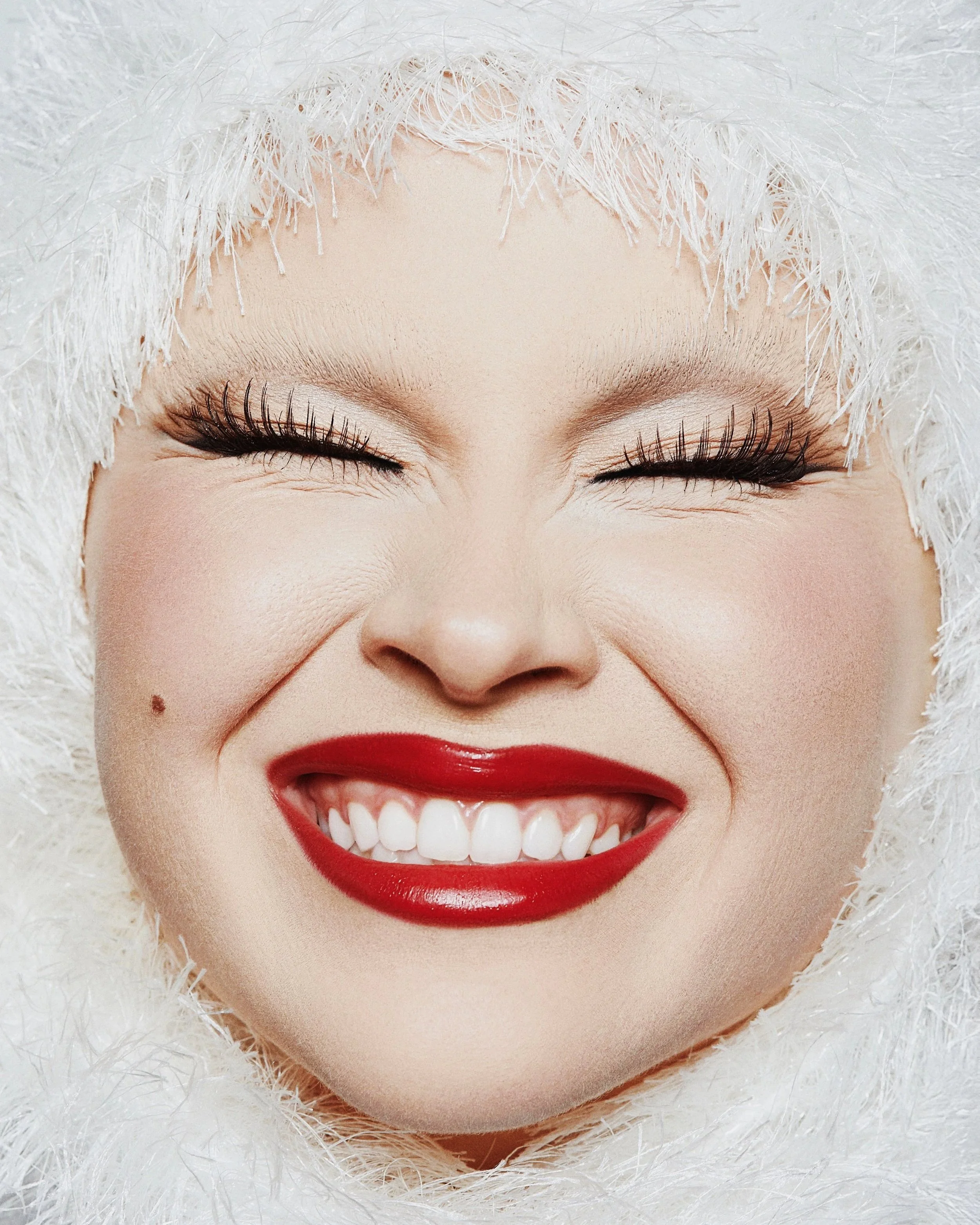 A smiling woman with red lipstick, closed eyes, and fair skin, surrounded by a fluffy white texture.