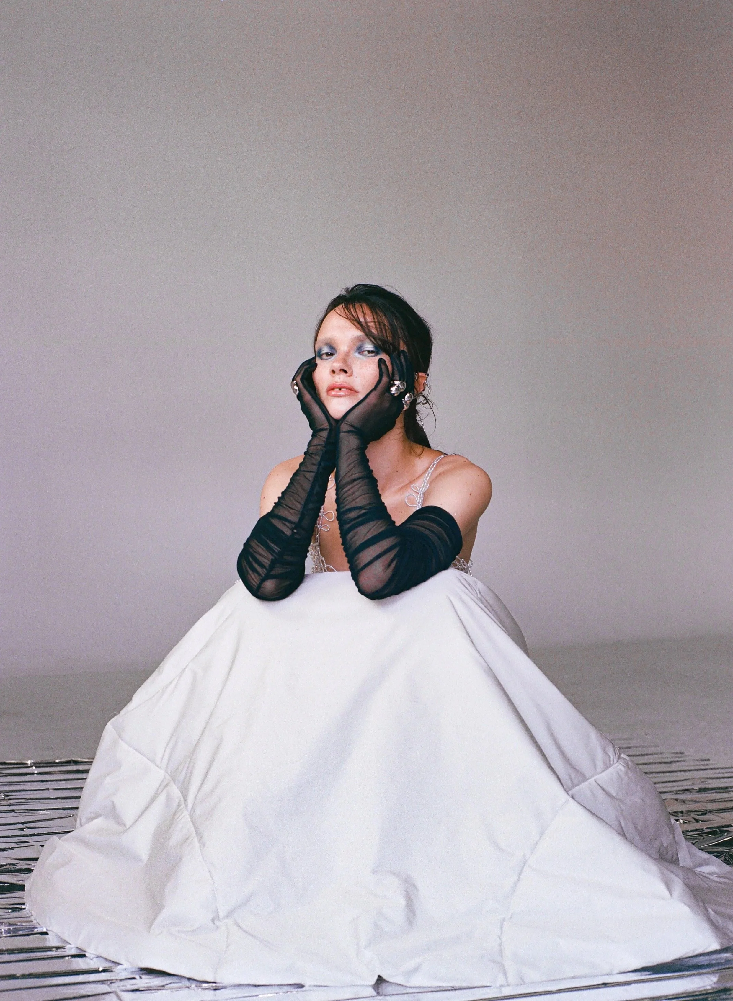 A person with black hair in a dark hairstyle, makeup, and jewelry, wearing a white wedding dress and long black sheer gloves, sits on a reflective surface with a plain background.
