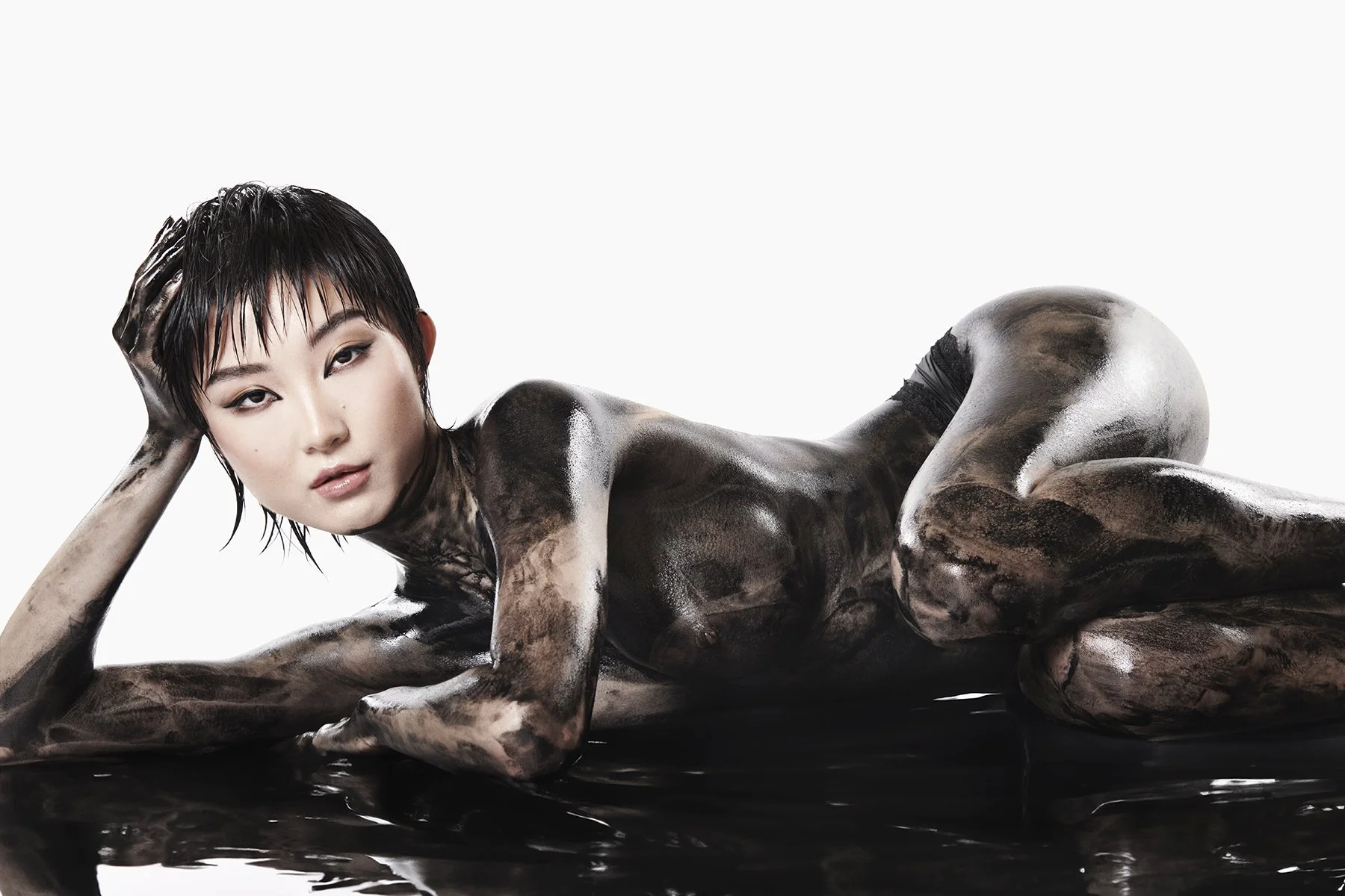 A woman with short black hair lying on a reflective black surface, covered in black and white paint, against a white background.