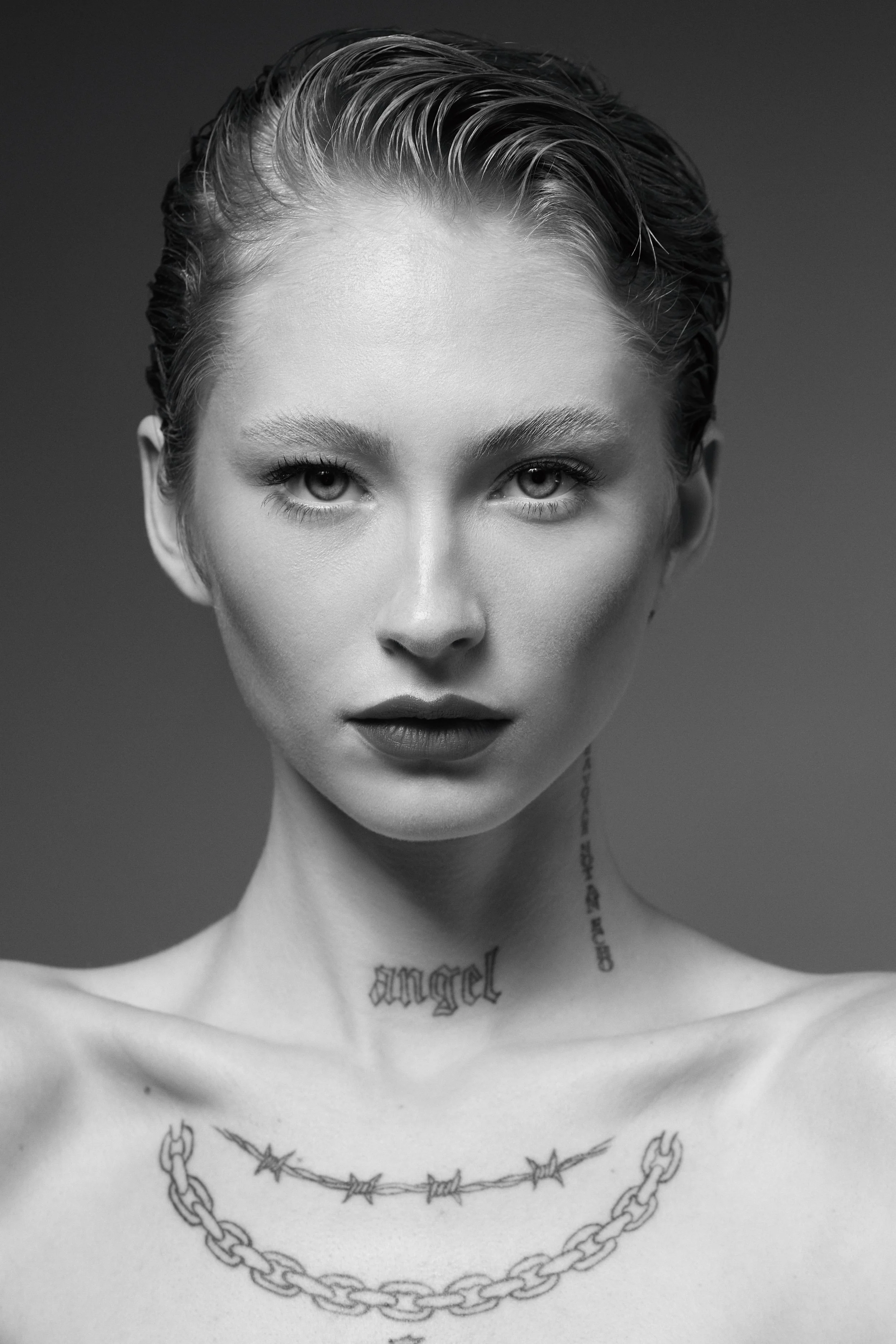 Black and white portrait of a young woman with short, slicked-back hair, wearing layered chains around her neck, with visible tattoos on her neck and chest