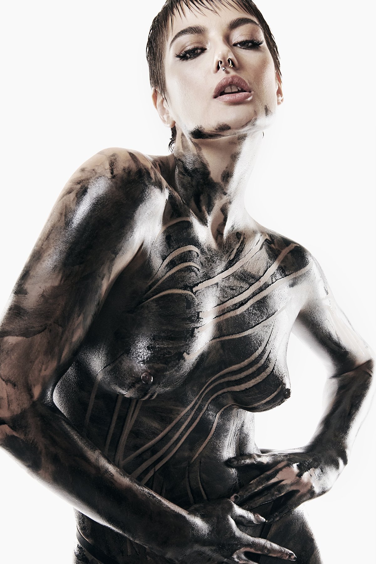 A woman with short hair and makeup, including eyeliner and lipstick, has black and white paint or ink tattoos covering her chest, arms, neck, and face, creating an abstract design against a white background.