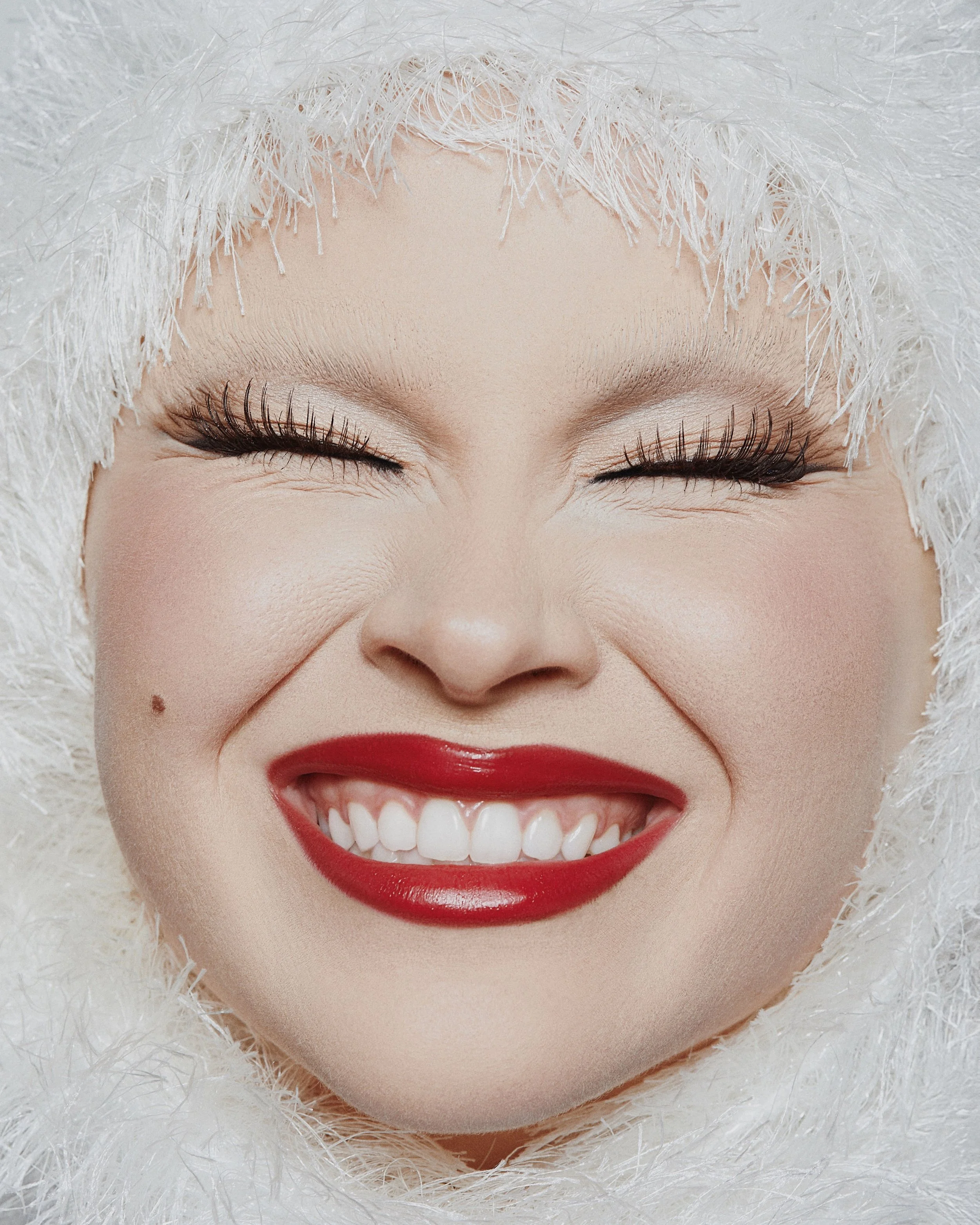 A woman smiling with eyes closed, wearing red lipstick, with a white, fluffy hood or fur around her face.