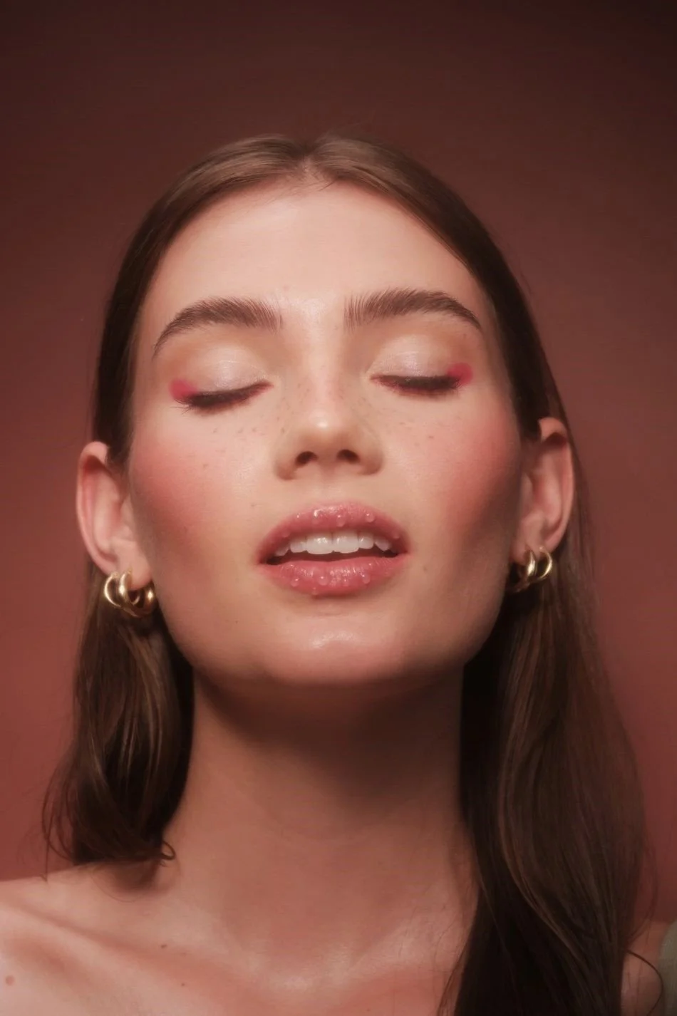 A young woman with fair skin, closed eyes, and brown hair, wearing gold hoop earrings, and glossy lips, with a neutral background.