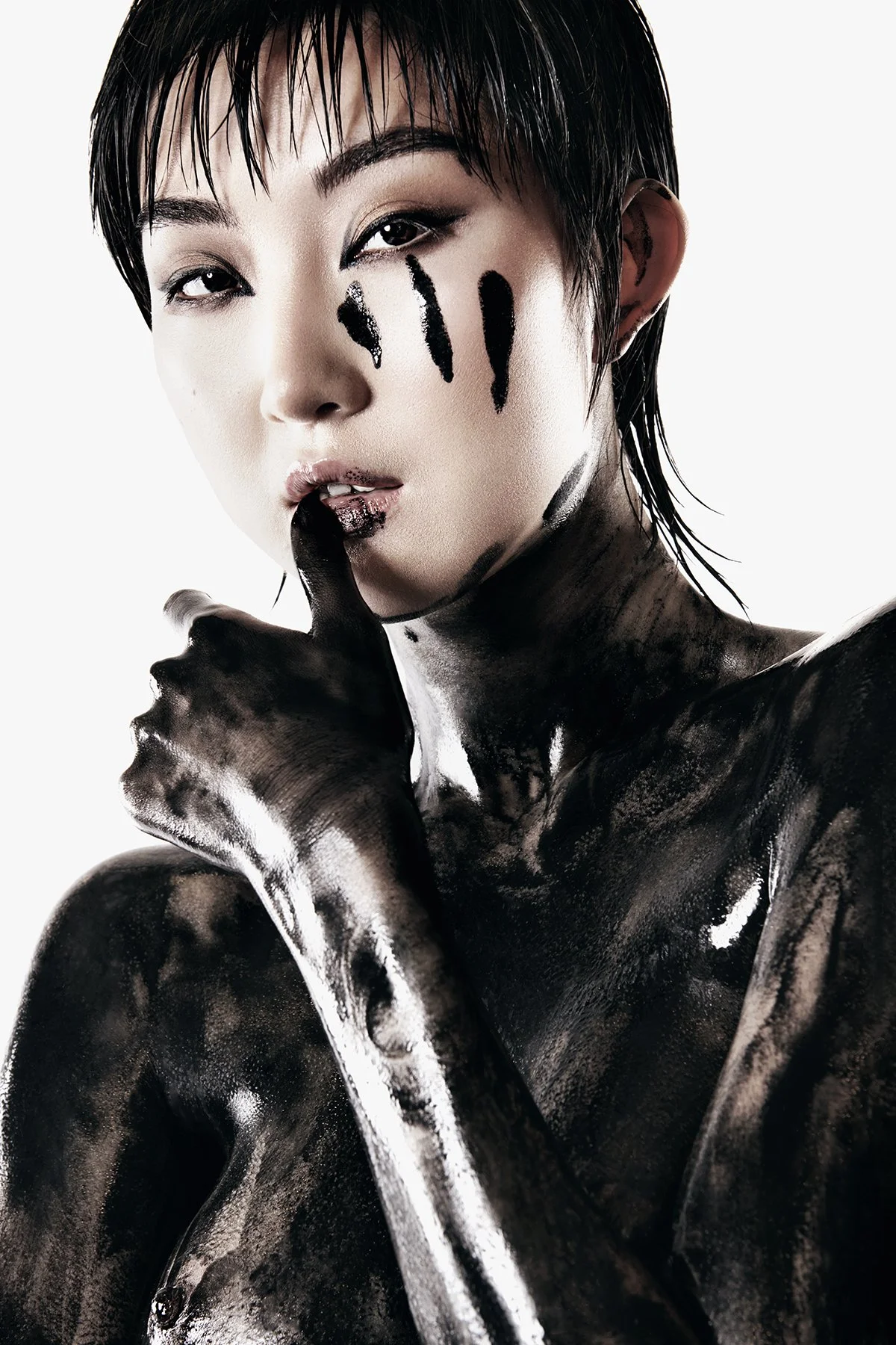 A woman with short dark hair, painted black and white on her body, touching her lips with gloved hand, with black paint streaks on her face.