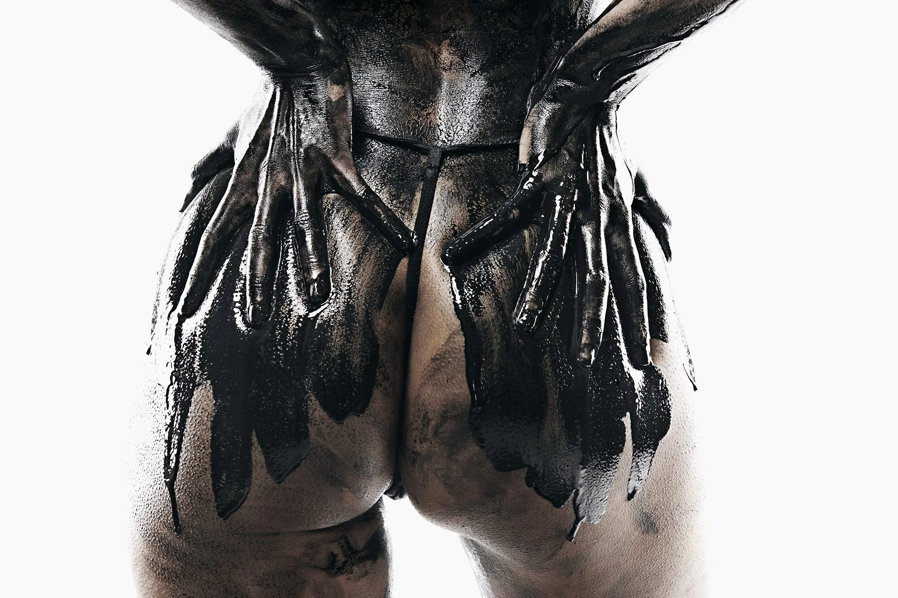 A person with dark skin is covered with black paint or a similar substance, with the paint dripping down their body. The person's hands are placed on their hips, and the focus is on their back and lower body against a white background.