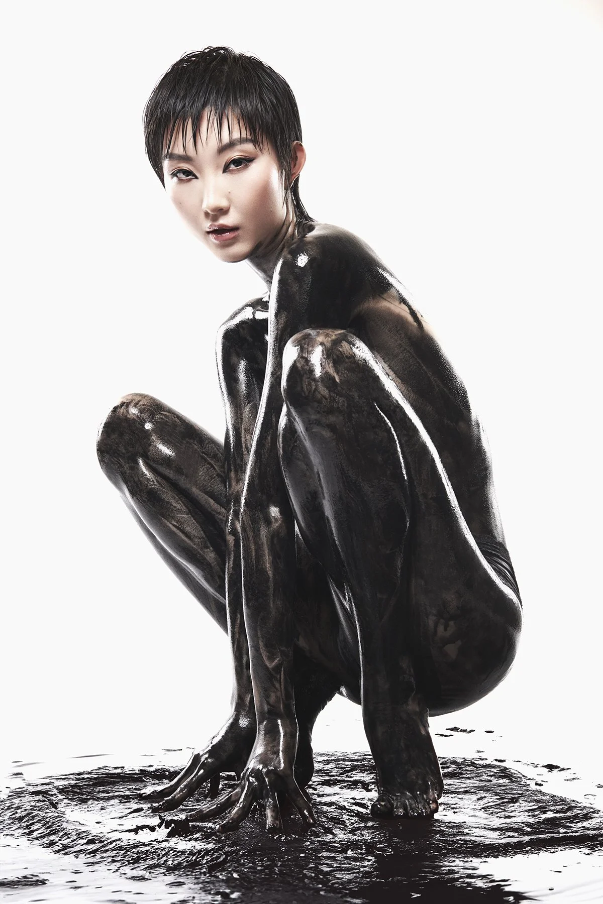 A woman in a crouching position with black, glossy body paint or material covering her body, against a white background.