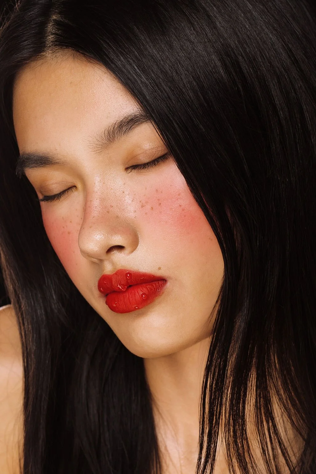 Close-up of a woman with dark hair, closed eyes, rosy cheeks, and red lips.