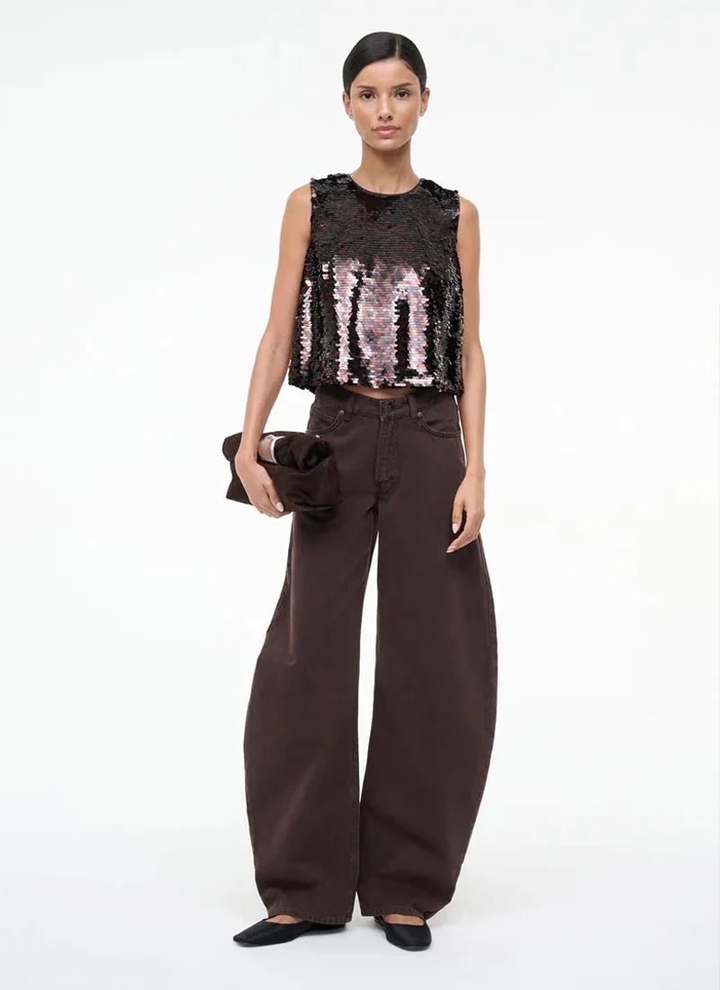 A woman with dark hair, wearing a sleeveless, black sequined top, loose dark brown pants, holding a small brown clutch, and wearing black flats, standing against a plain white background.