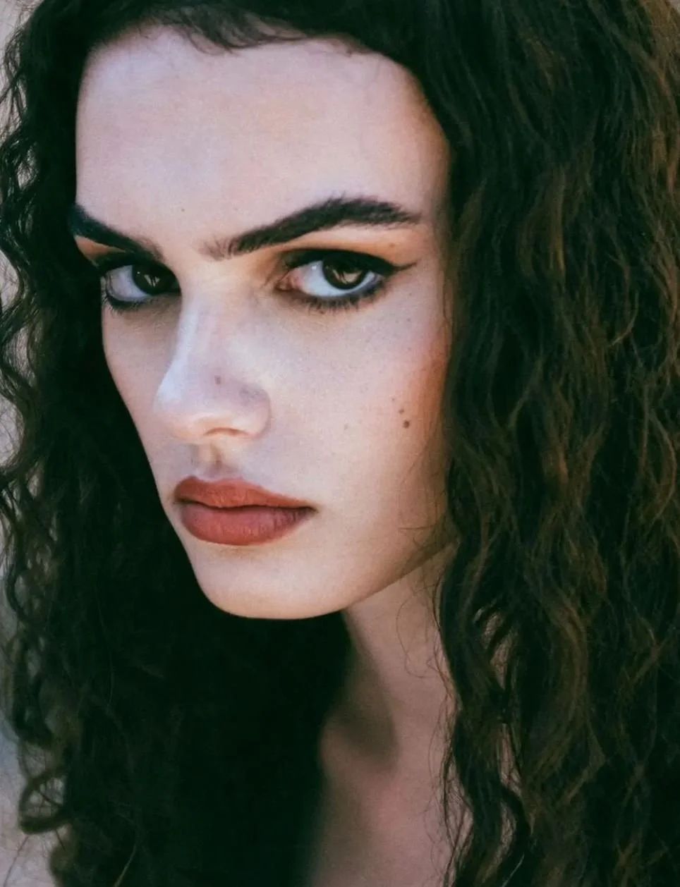 Close-up of a woman with dark curly hair, green eyes, and light skin, wearing makeup including eyeliner and lipstick.