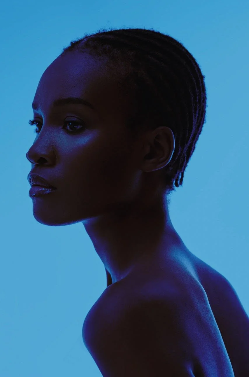 Side profile of a woman with short dreadlocks, illuminated by blue lighting, against a blue background. Dark skin makeup black woman makeup artist, colormatching, brown girls, mali magic, editorial, vogue