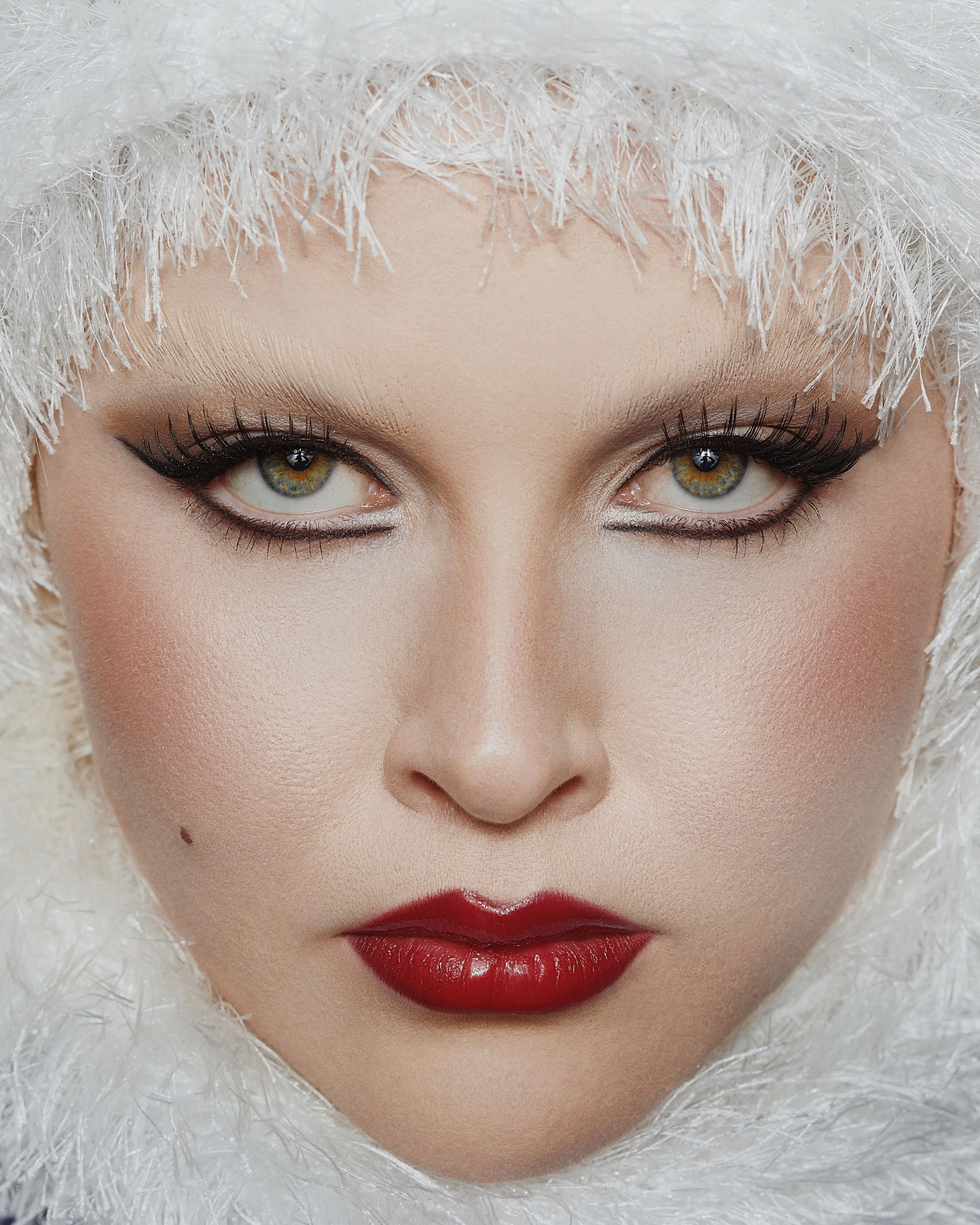 Close-up of a woman's face with bold makeup, red lips, and a white feathered hood.