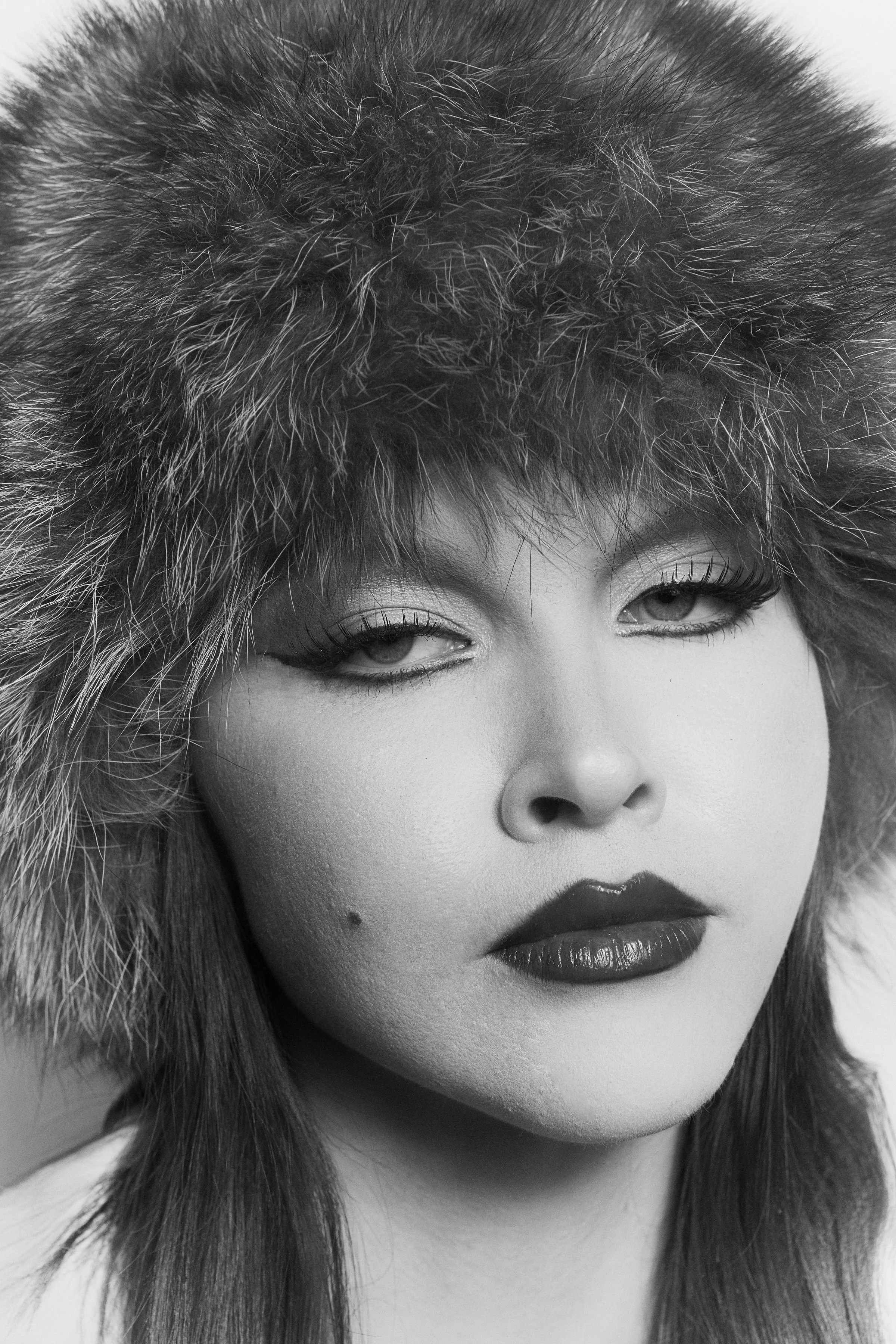 A black-and-white close-up of a woman wearing a furry hat, with makeup and long hair.