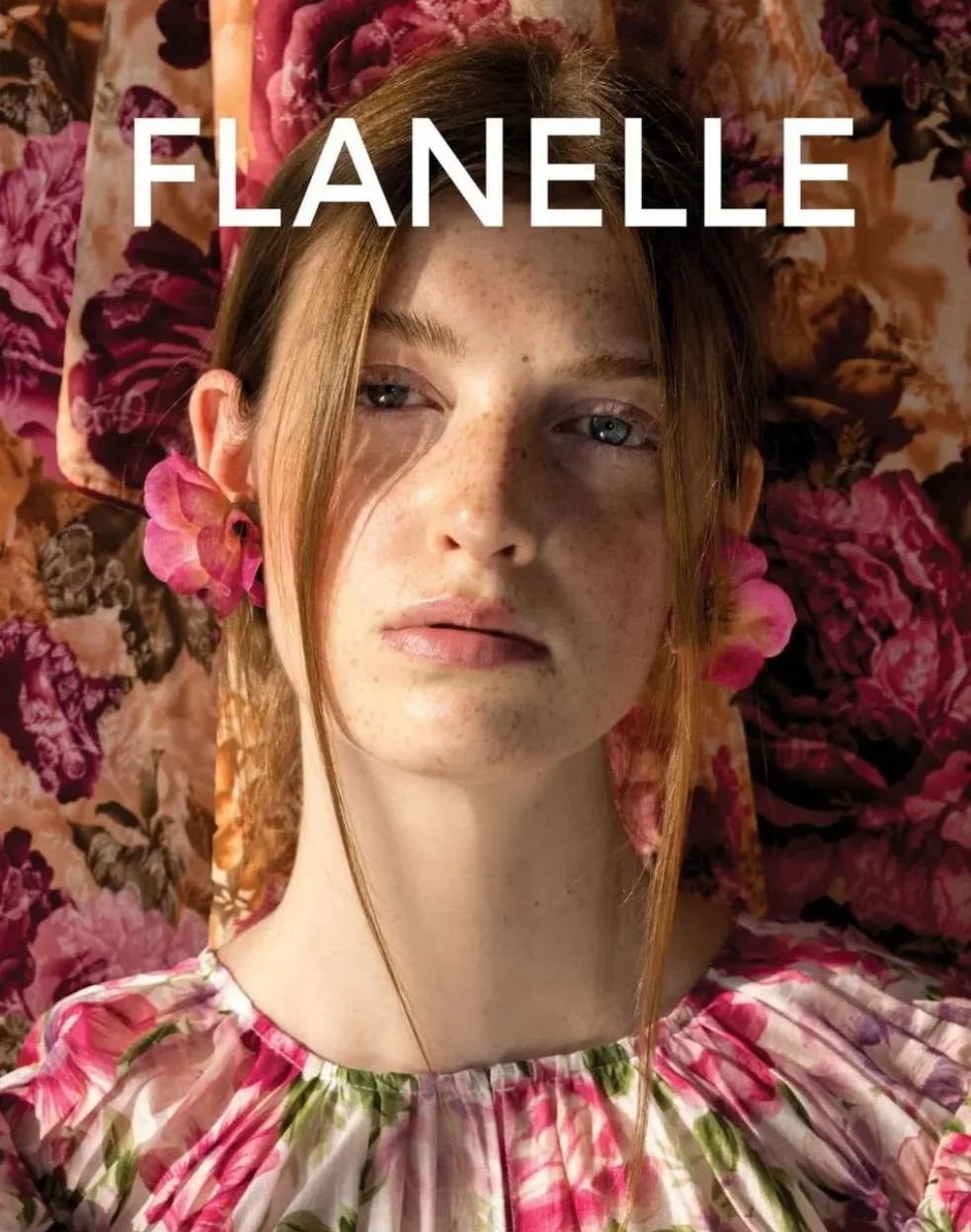 A young woman with freckles and blue eyes lying among pink and purple flowers, wearing a pink floral dress, with pink flowers in her hair and the name "Flanelle" written across the top. editorial fashion beauty toronto makeup clean 