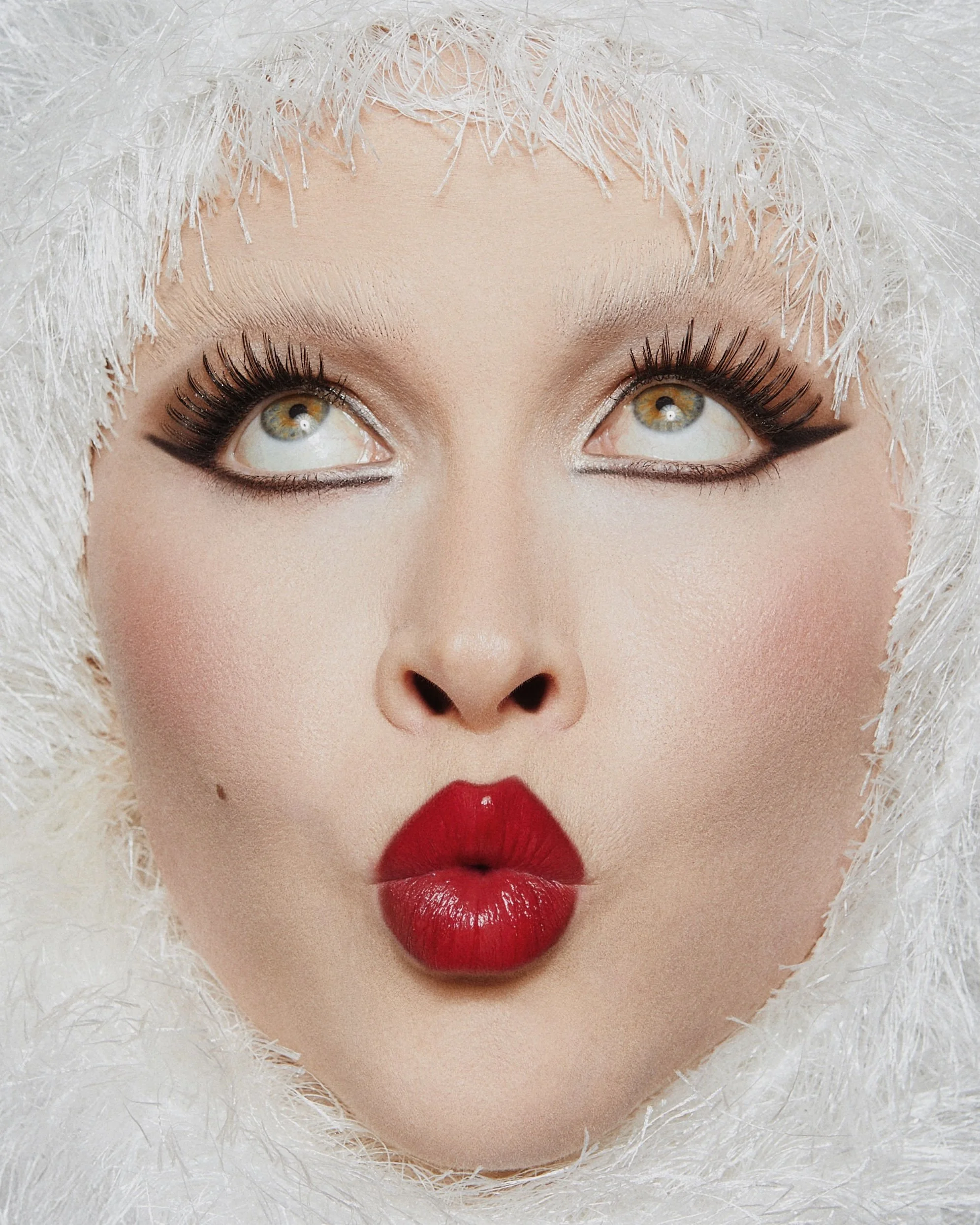 Close-up of a woman with photographed makeup, wearing a white furry hood, making a kissy face expression.