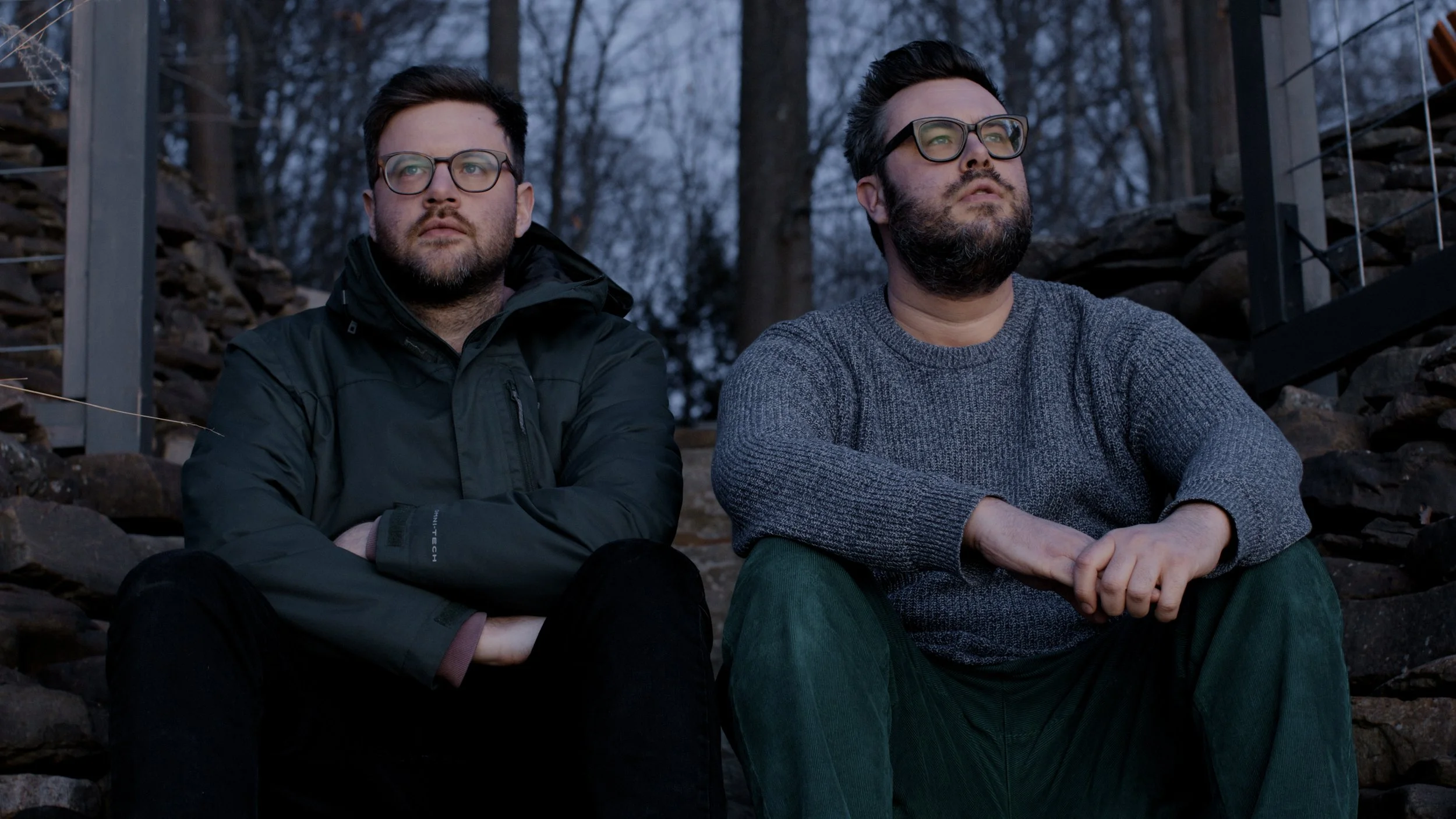 Two men sitting outdoors on stone steps, looking pensively into the distance, with trees and a wooden structure in the background.