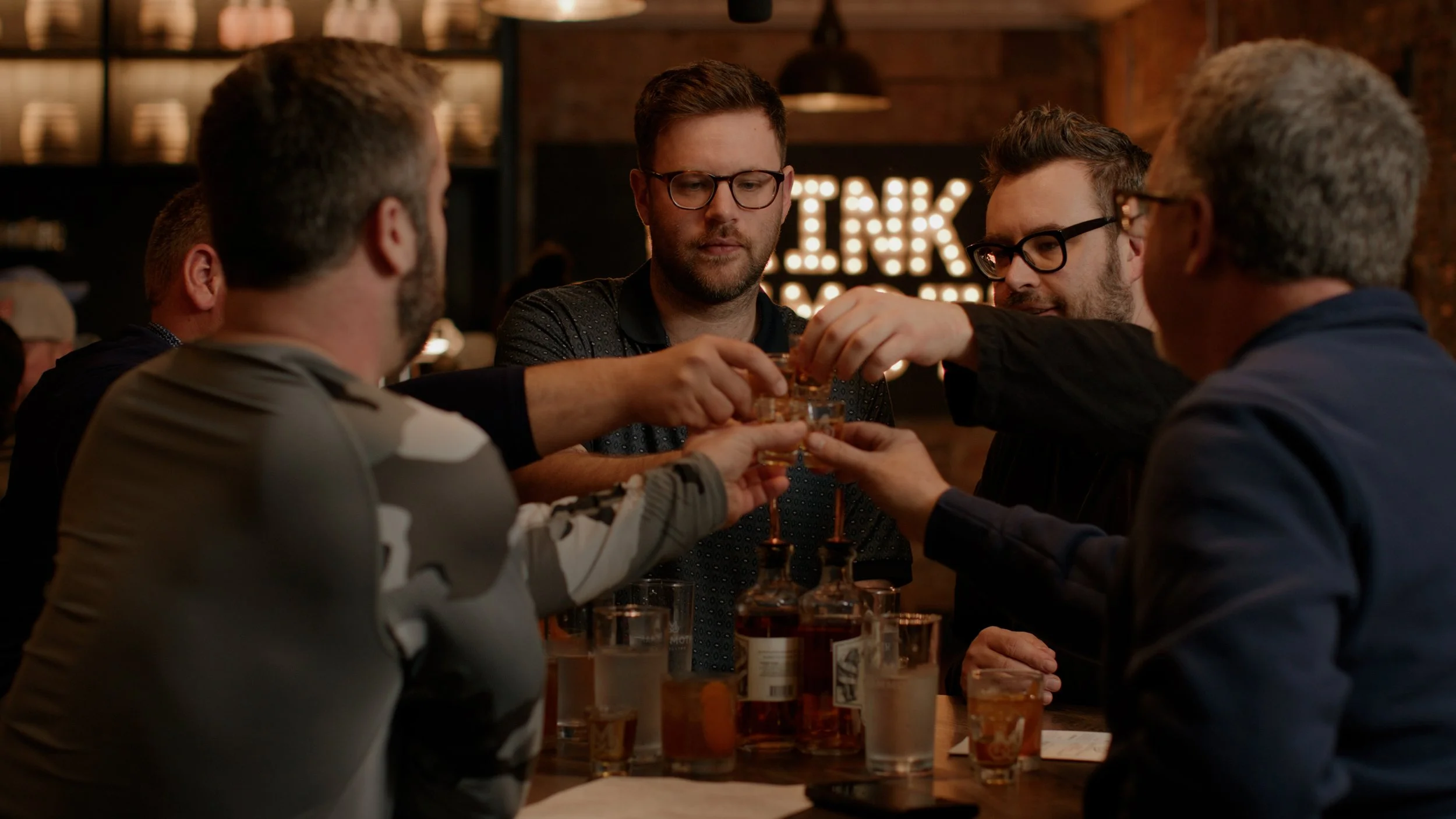 A group of five men sitting around a table at a bar, toasting with glasses of whiskey, with bottles and glasses on the table and a lit sign in the background.