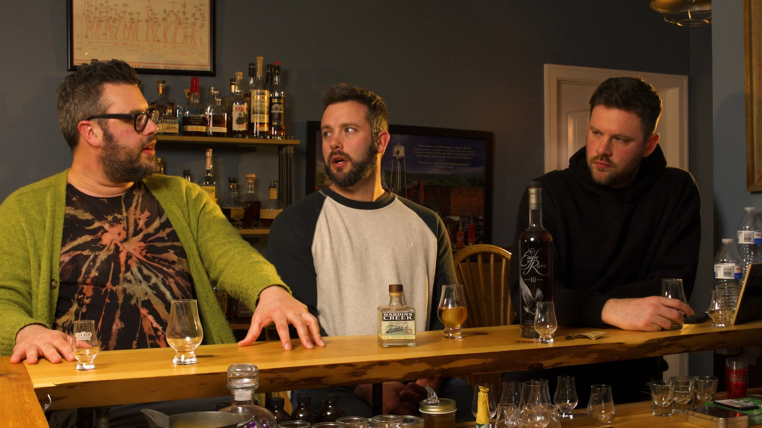 Three men sit at a bar having a conversation, with various bottles of liquor and glasses on the bar and shelves behind them.