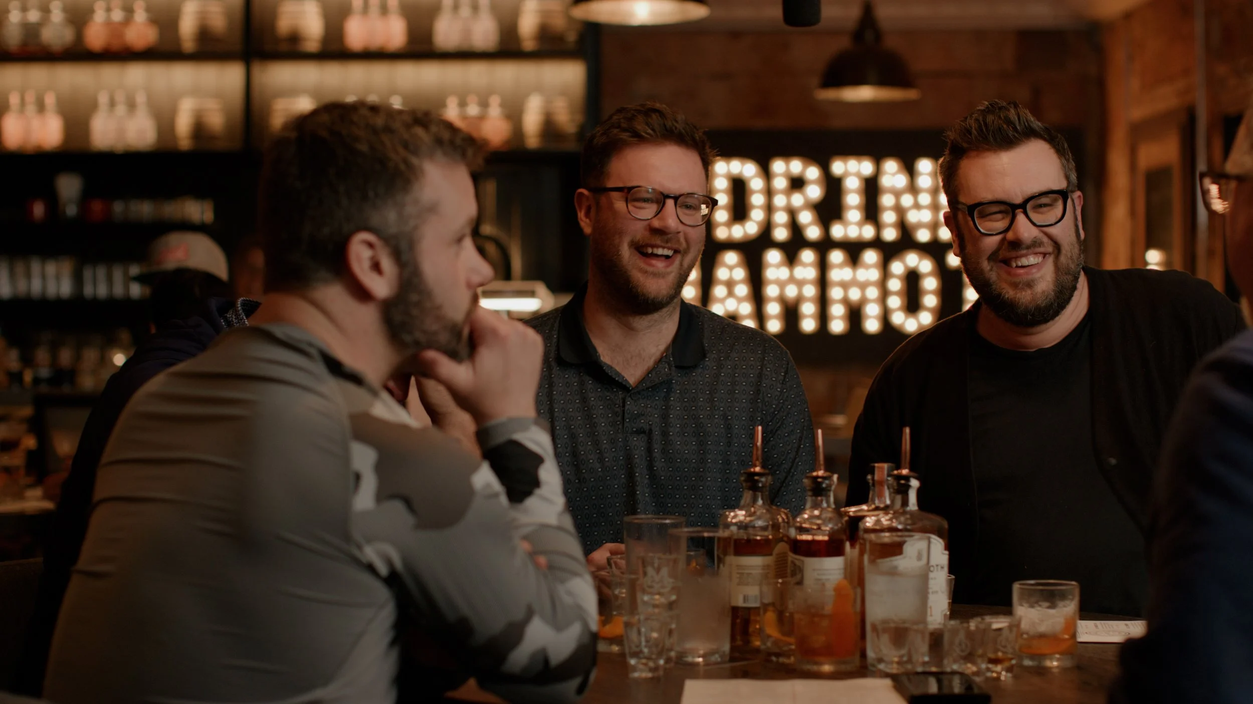 Three men sitting at a bar and laughing, drinks on the table, in a restaurant or bar with a sign that reads "Drinks & Ammo" in the background.