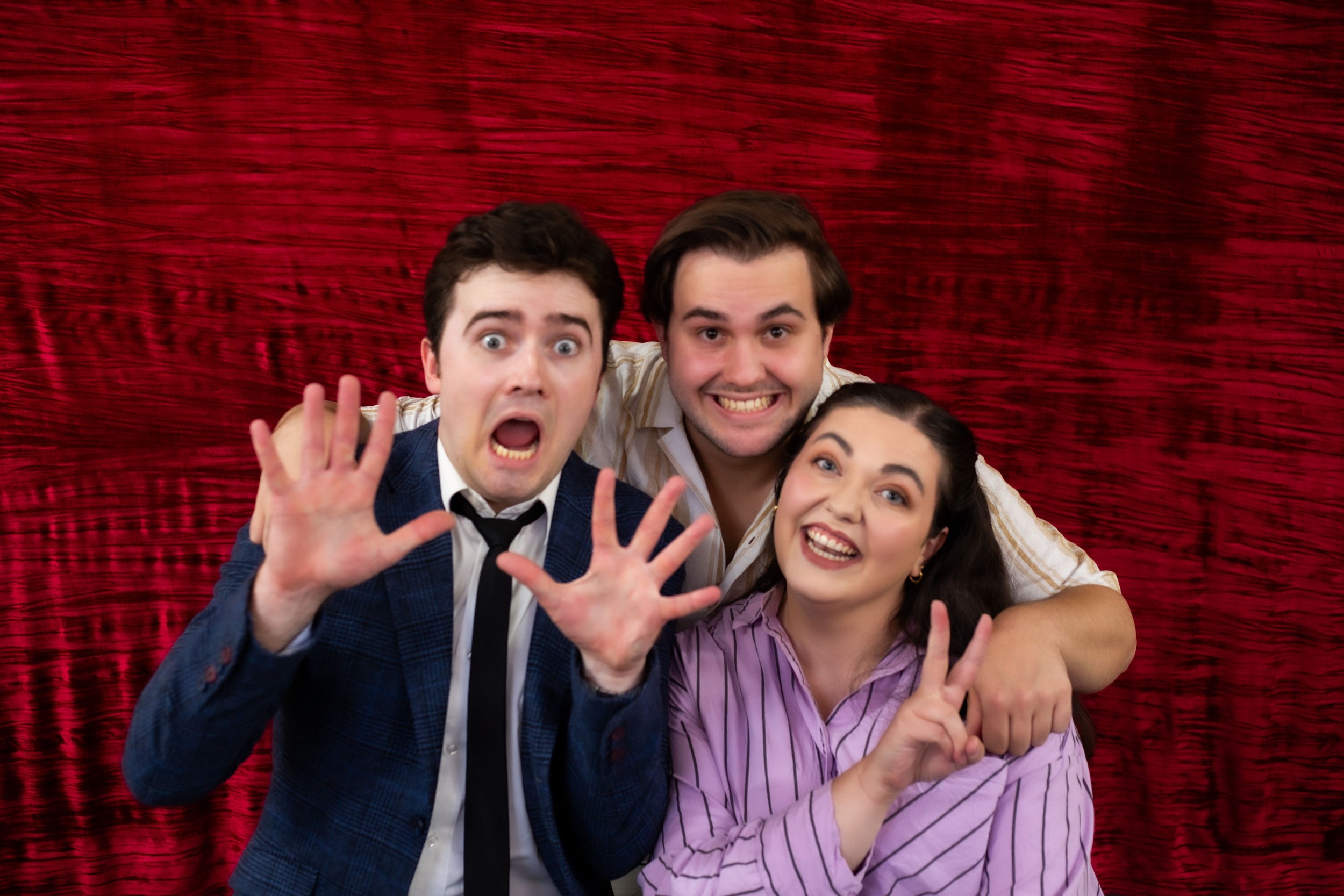 Three people posing playfully in front of a red curtain, displaying humorous expressions. One person in a blue suit and tie has hands raised, another is wearing a striped shirt, and the third in a striped blouse is making a peace sign.