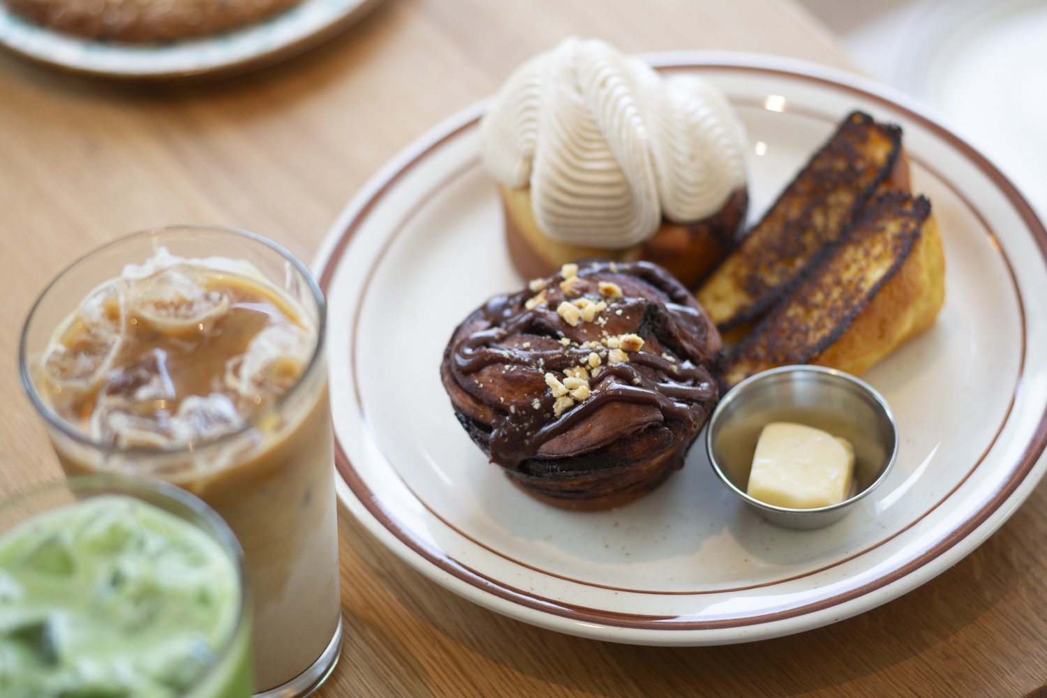 Plate with a cupcake topped with white frosting, two pieces of banana bread, a chocolate muffin with nuts and chocolate drizzle, and a small container of butter. Two glasses of iced coffee and a green drink are partially visible in the foreground.