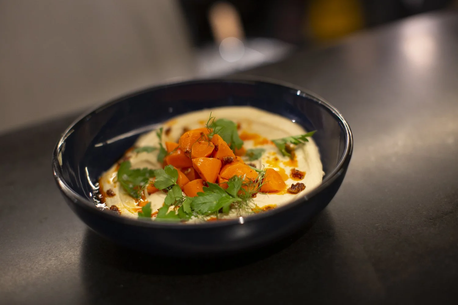 A black bowl with a creamy spread topped with diced orange carrots, fresh green herbs, and small dark spices, garnished with cilantro leaves.