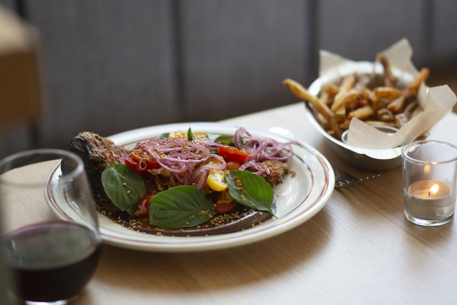 A plate of fish with colorful vegetables and herbs, a side of fries, a glass of wine, and a lit candle on a wooden table.