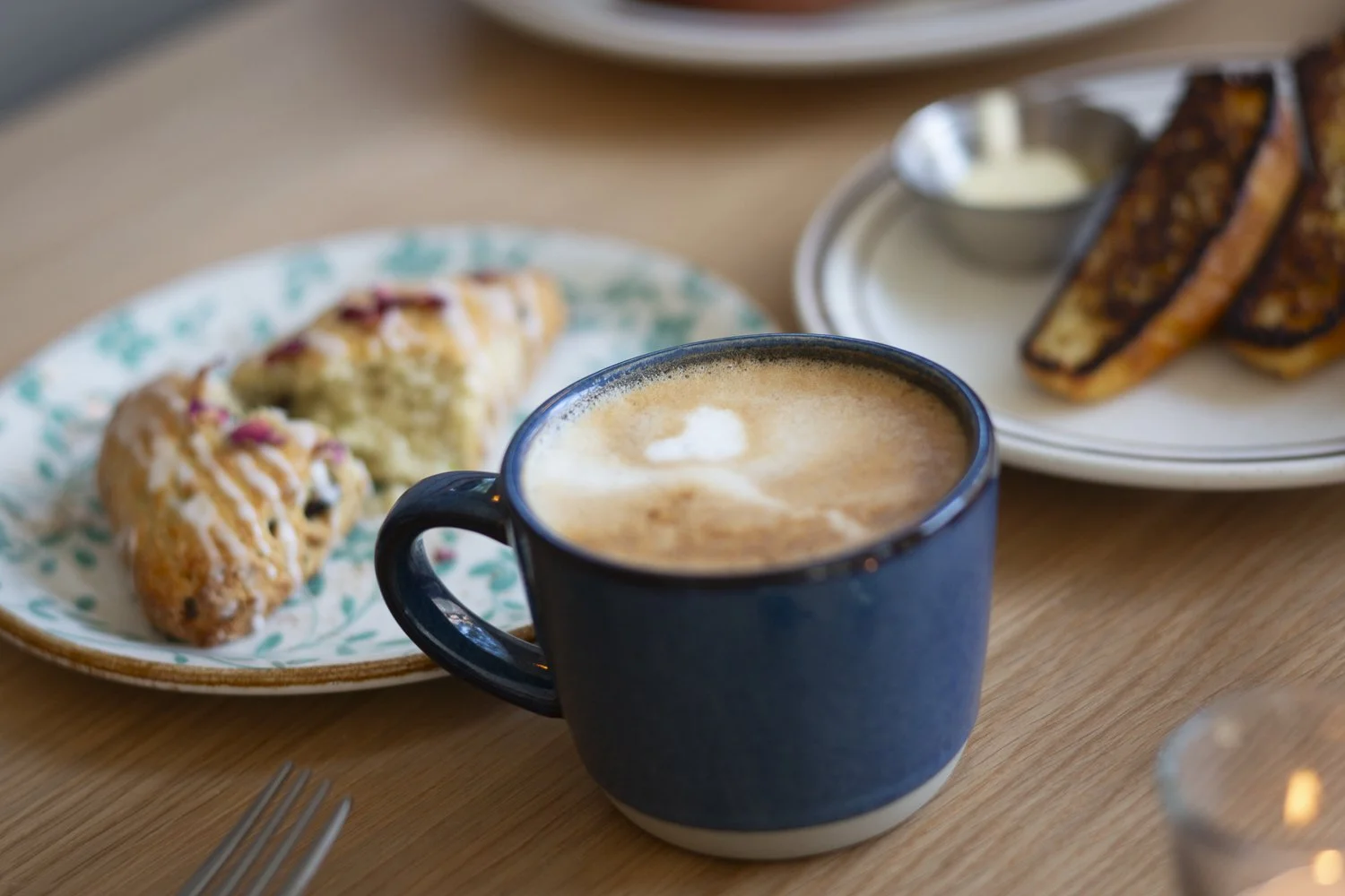 A blue mug filled with a frothy cappuccino on a wooden table. In the background, a white plate holds two scones with pink drizzle, and another plate has two sausage breakfast sandwiches with a small bowl of white dipping sauce.