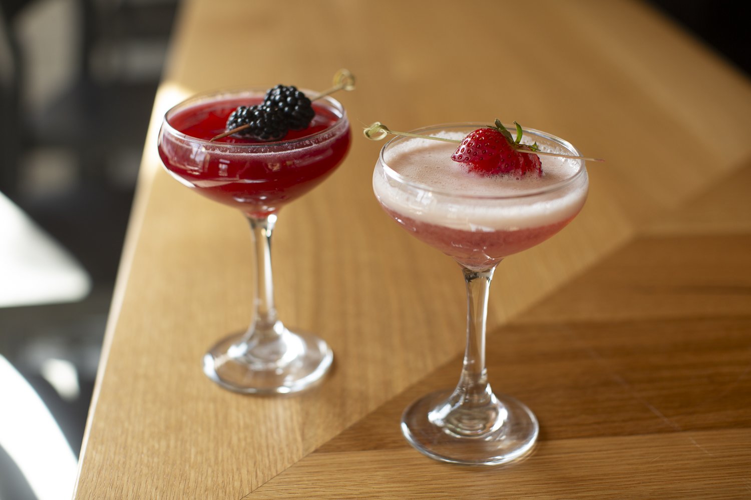 Two cocktail glasses with pink and red layered drinks, garnished with strawberries and blackberries on skewers, placed on a wooden surface.