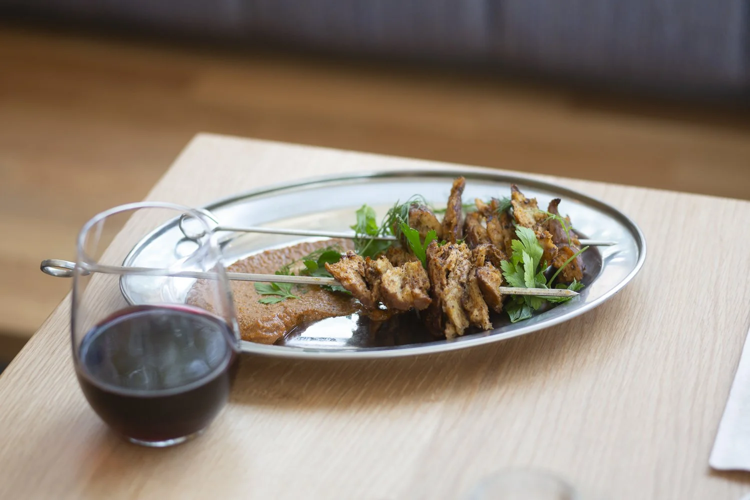 A silver oval platter with grilled meat skewers garnished with green herbs, accompanied by a side of sauce or chutney. A glass of red wine is placed next to the platter. The items are on a light wooden table.