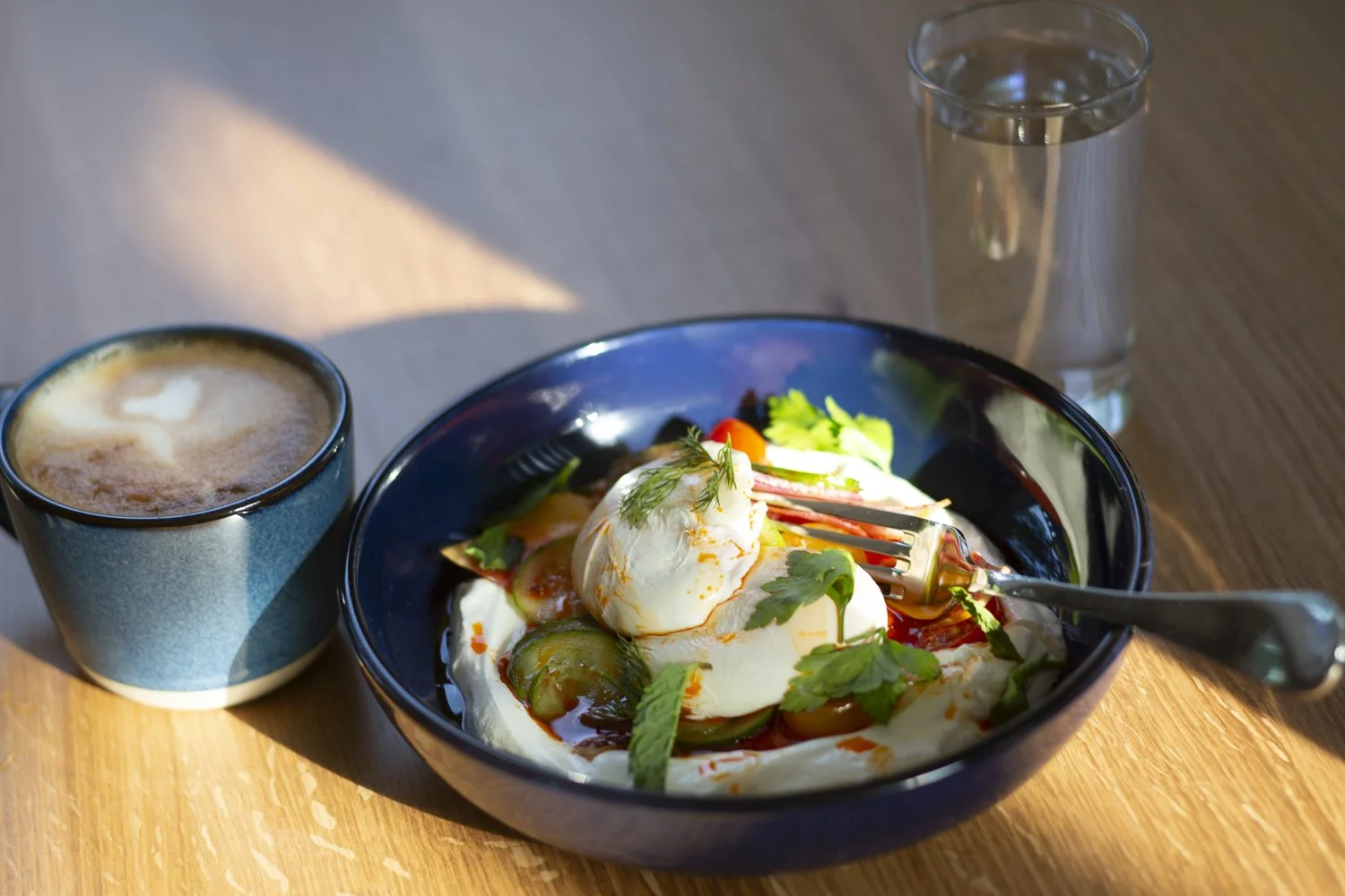 A breakfast consisting of a bowl of poached eggs, sliced cucumbers, cherry tomatoes, and fresh herbs, with a cup of coffee and a glass of water on a wooden table.