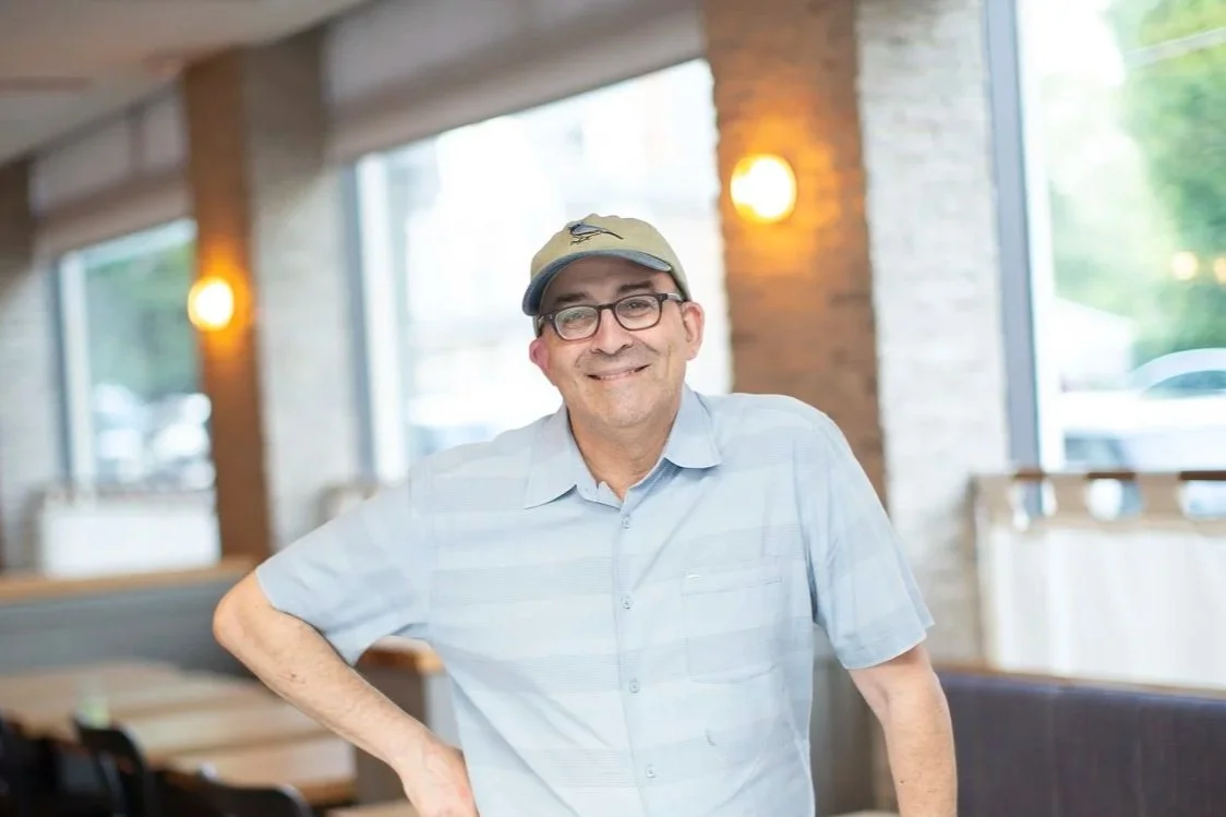 Owner, Fred Mogul, wearing glasses, a cap, and a light blue striped shirt stands inside the Blue Warbler restaurant with exposed brick walls and large windows.