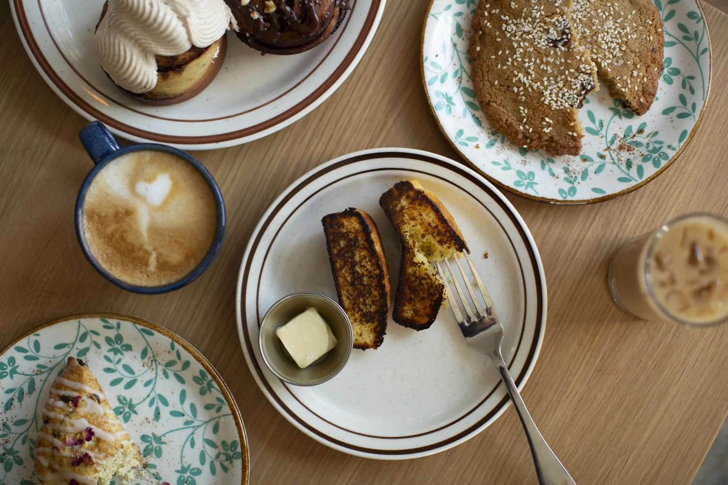 Assorted desserts and beverages on a wooden table, including a slice of cinnamon toast with butter, a chocolate chip cookie, a small cake with icing, a pastry topped with white icing and pink sprinkles, a cup of coffee with frothy milk, and an iced c