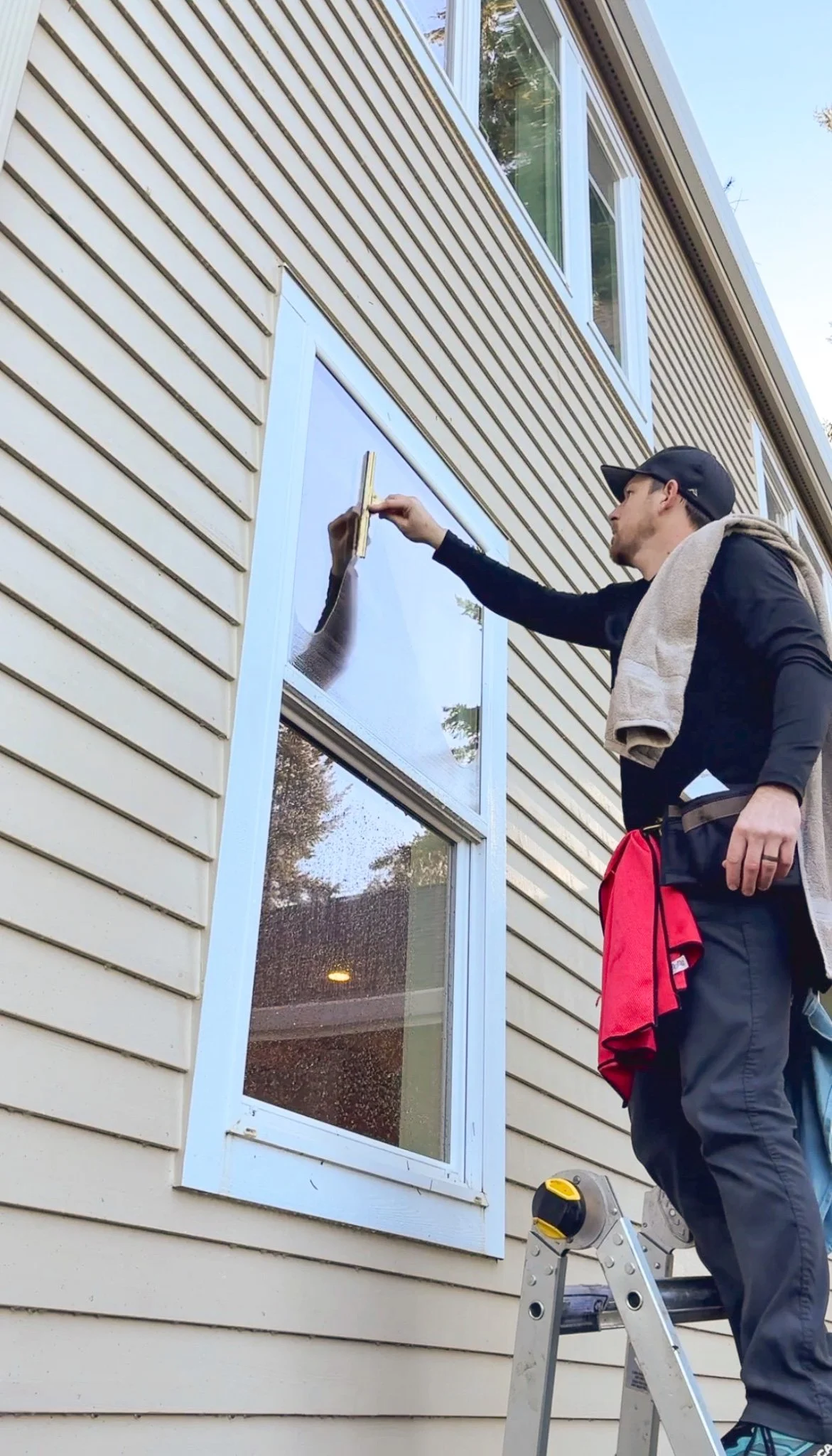 A man standing on a ladder cleaning the exterior window of a house with a squeegee.